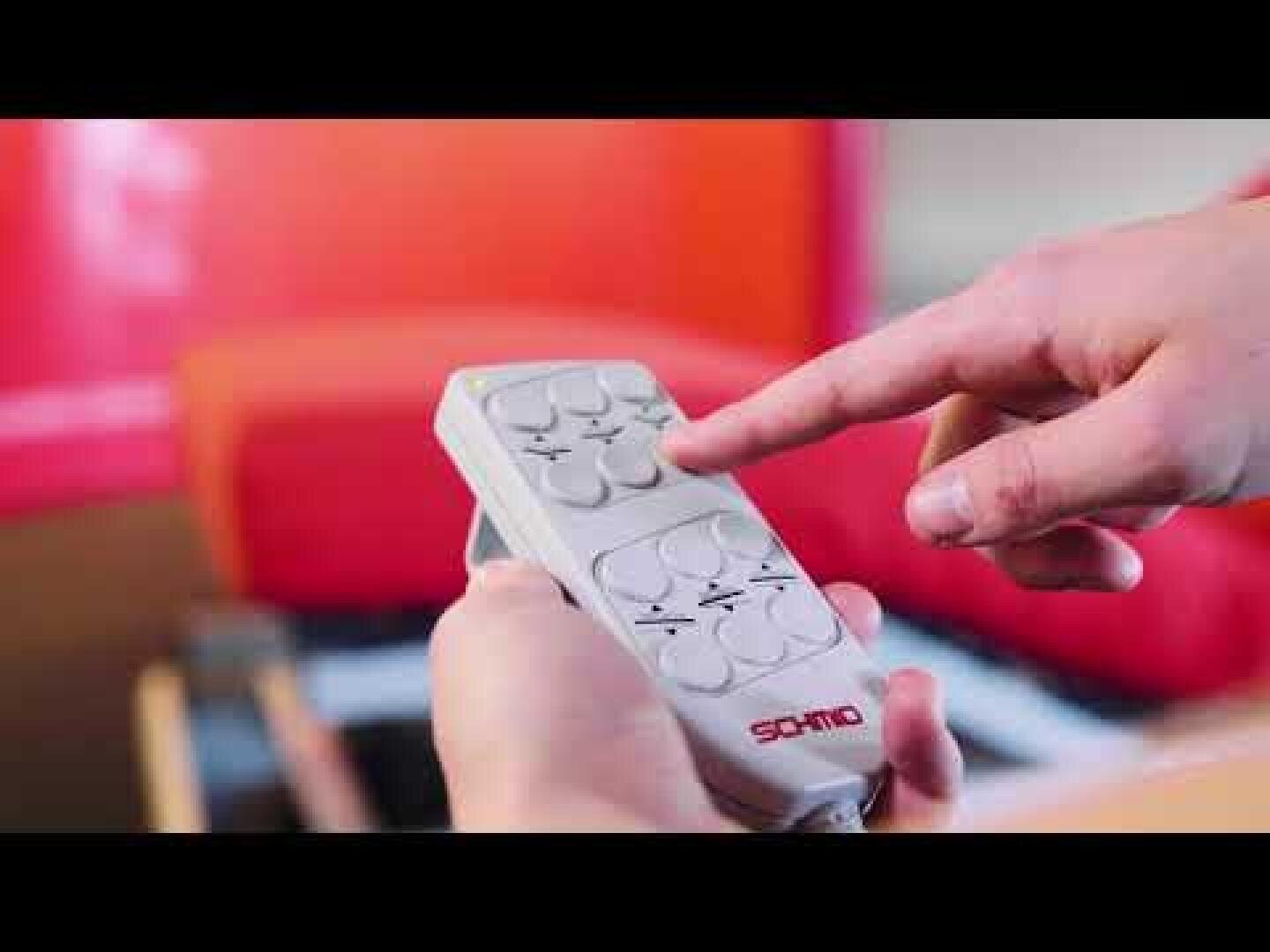 Eine Person hält eine weiße SOMNO-Fernbedienung in der Hand und drückt eine Taste, während im Hintergrund ein verschwommener roter Stuhl zu sehen ist.