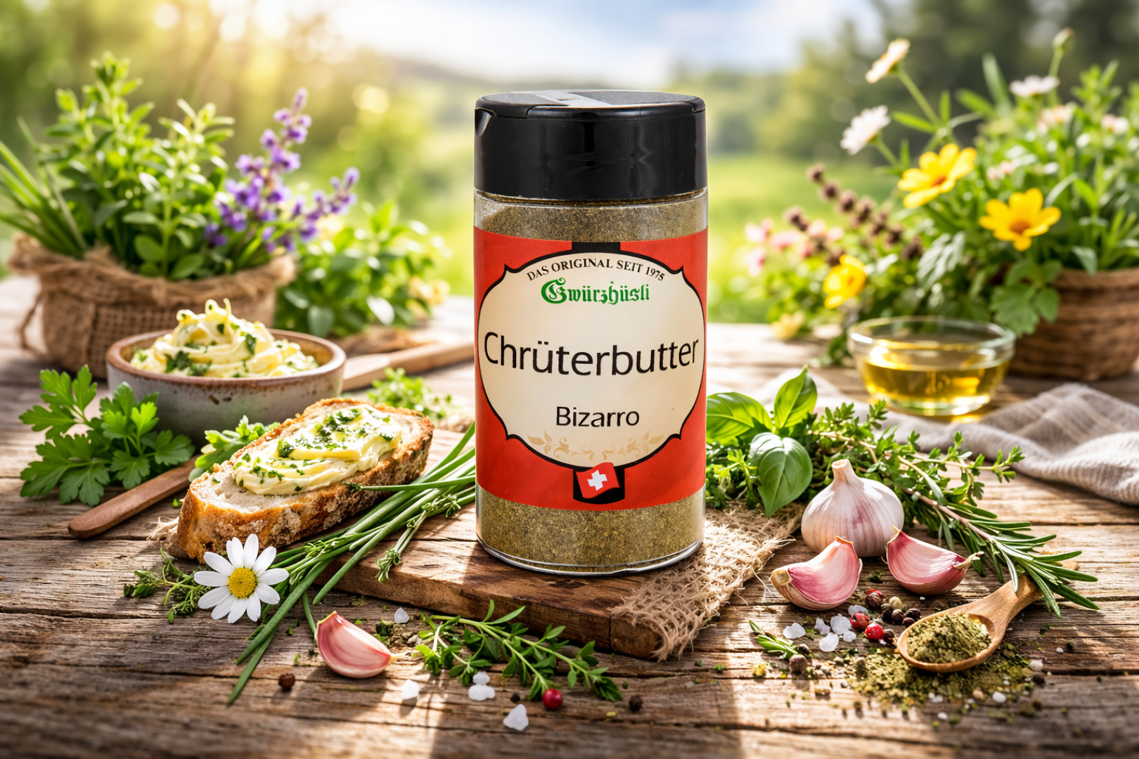 Ein Behälter mit der Gewürzmischung Chrüterbutter steht auf einem rustikalen Holztisch, umgeben von frischen Kräutern, Knoblauch, Blumen, Brot mit Kräuterbutter und einem Schälchen Olivenöl, im Hintergrund eine malerische grüne Landschaft.