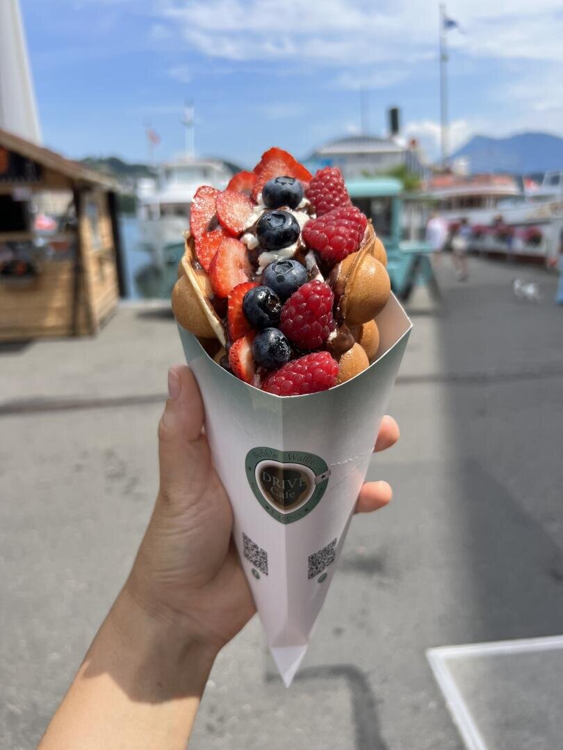 Eine Hand hält eine Schaumwaffel mit Schlagsahne, Erdbeeren, Heidelbeeren und Himbeeren, die in einer Papiertüte serviert wird. Die Essensstände im Freien und die Berge sind im Hintergrund verschwommen.