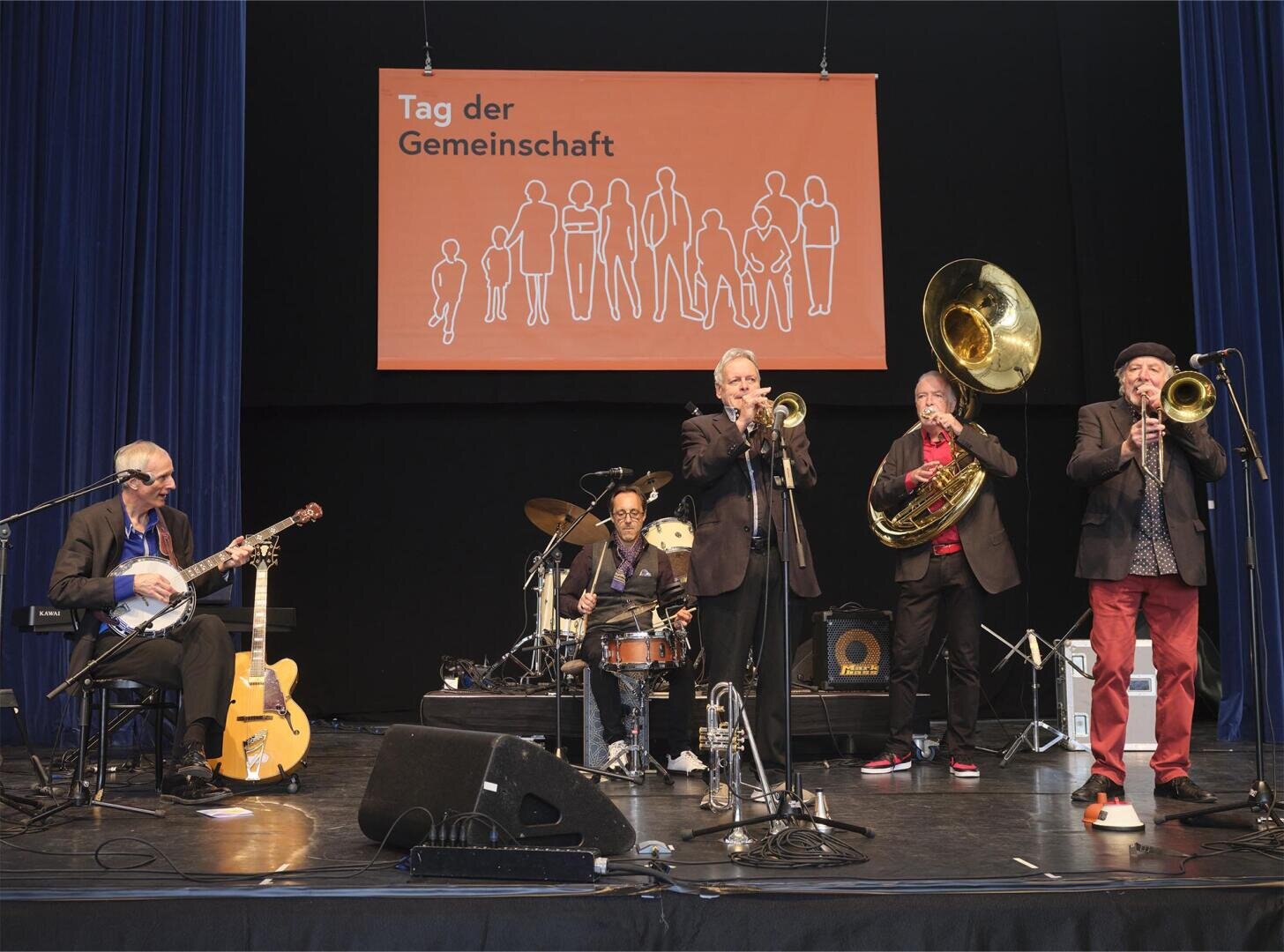 Auf der Bühne tritt eine fünfköpfige Band mit Gitarre, Schlagzeug, Posaune, Sousaphon und Banjo auf. Hinter ihnen hängt ein orangefarbenes Transparent mit der Aufschrift "Tag der Gemeinschaft" und den Umrissen von Menschen.