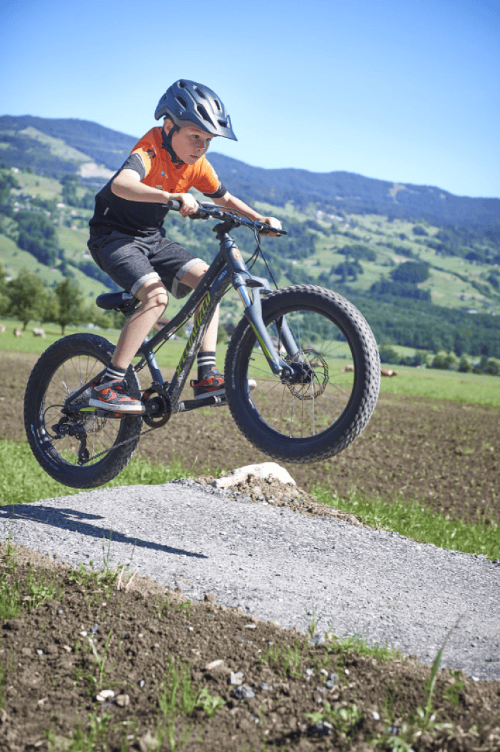 Ein kleiner Junge mit Helm und orangefarbenem Hemd fährt mit seinem Mountainbike über einen kleinen Sprung auf einem Kiesweg, mit grünen Hügeln und blauem Himmel im Hintergrund.