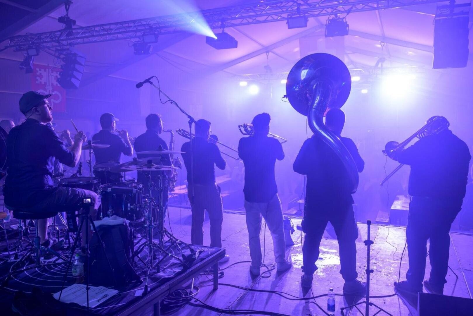 Eine Blaskapelle tritt auf einer schwach beleuchteten Bühne mit blauen und violetten Lichtern auf, von hinten gesehen. Die Musiker spielen verschiedene Blechblasinstrumente und Trommeln und blicken auf ein Publikum, das von Nebel und hellen Bühnenlichtern verdunkelt wird.