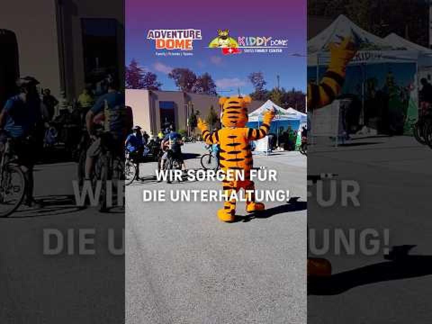Eine Person in einem Tigerkostüm unterhält die Leute bei einer Freiluftveranstaltung mit Radfahrern und Zelten im Hintergrund. Der deutsche Text auf dem Bild lautet Wir sorgen für die Unterhaltung.