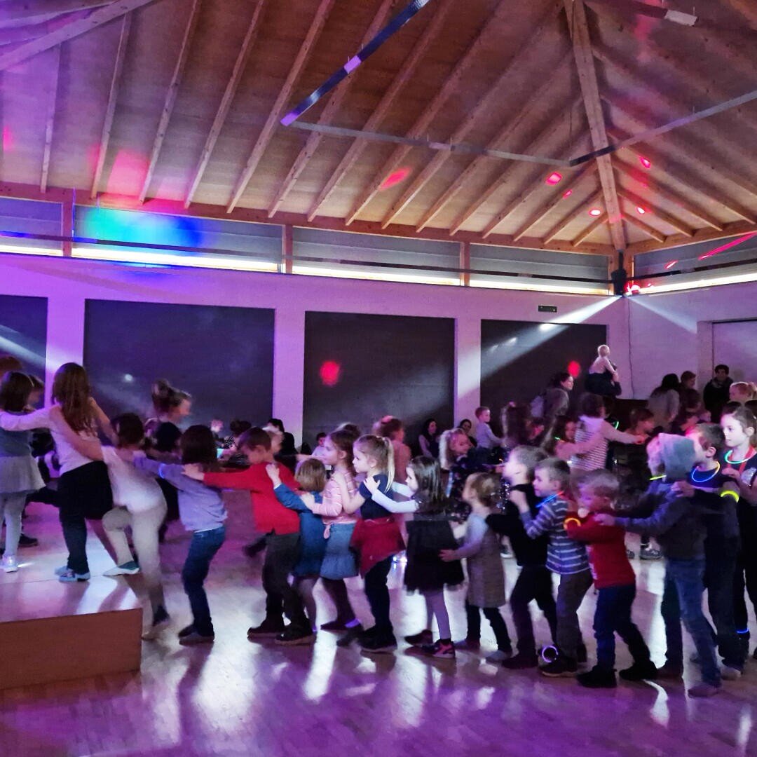 Eine Gruppe von Kindern bildet eine Conga-Linie und tanzt in einer Halle mit Holzdecken und bunten Partylichtern. Im Hintergrund sind weitere Kinder und Erwachsene zu sehen, die sich an dem lebhaften Ereignis erfreuen.