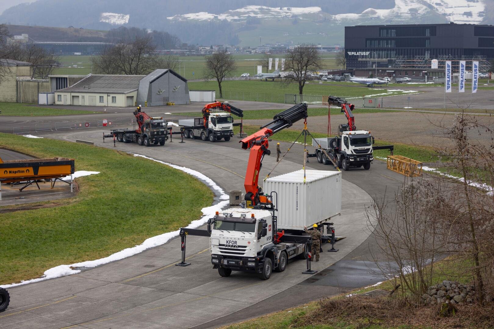 Vier Lastwagen mit großen orangefarbenen Kränen heben und transportieren weiße Schiffscontainer auf einer asphaltierten Straße in einem Industriegebiet, mit Schneefeldern, Grasflächen und Gebäuden im Hintergrund.
