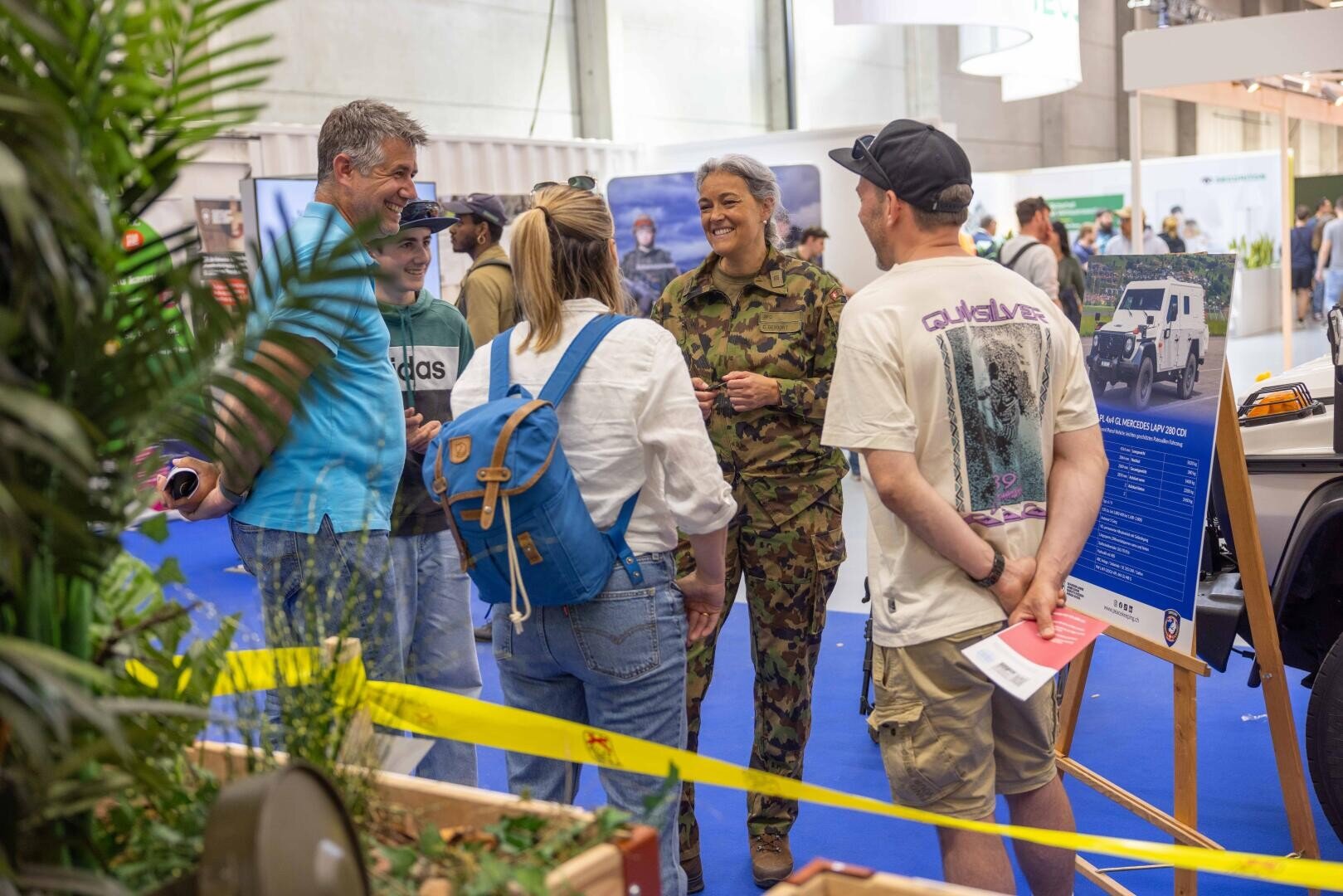 Eine Gruppe von Personen, darunter eine Frau in militärischer Tarnkleidung, steht auf einer Indoor-Ausstellung und unterhält sich. Im Hintergrund sind Informationsdisplays, Pflanzen und gelbes Klebeband zu sehen. Die Atmosphäre wirkt freundlich und einnehmend.