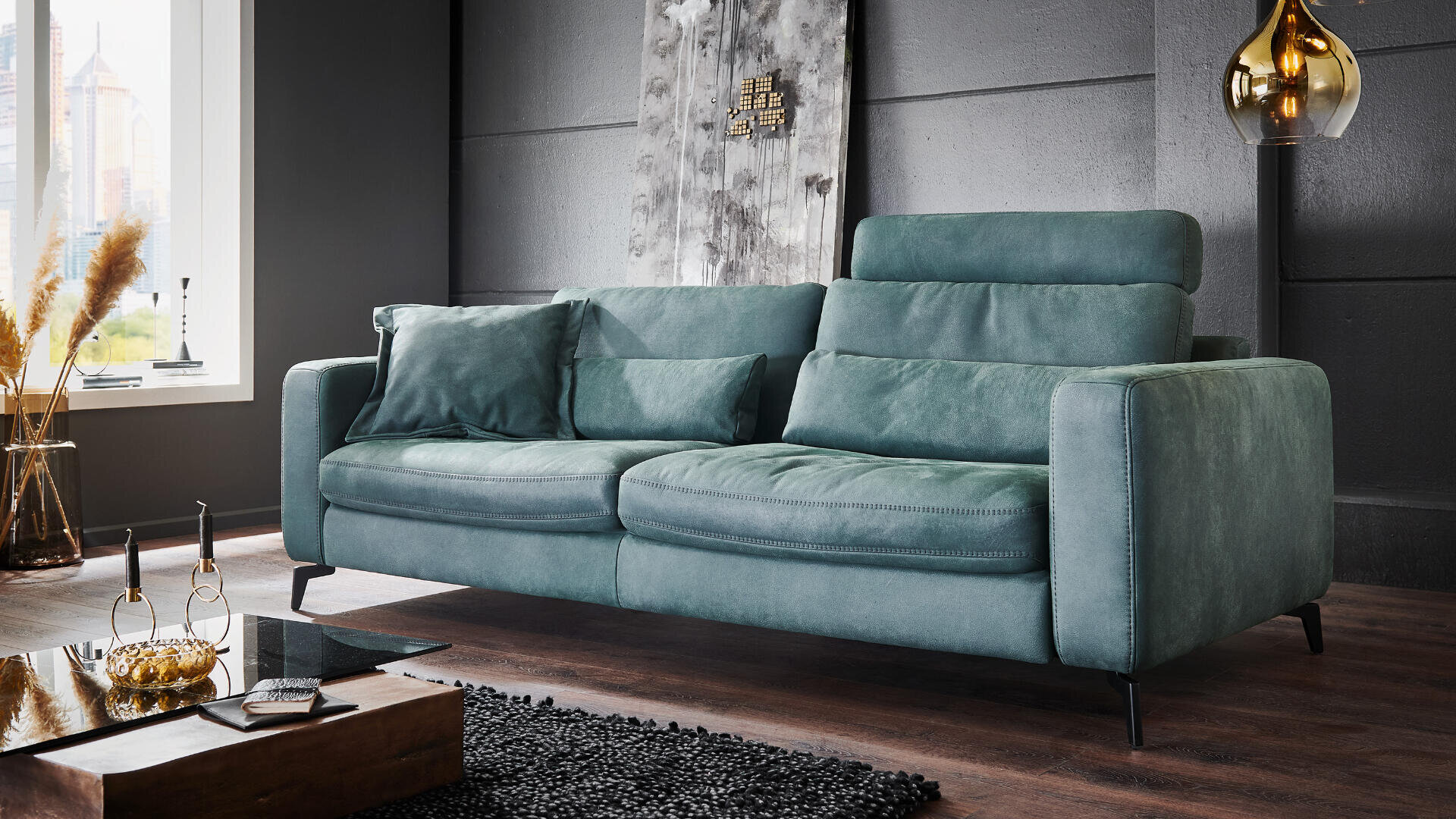 Ein modernes Sofa in Blaugrün mit weichen Kissen in einem stilvollen Wohnzimmer mit dunklen Wänden, einem Couchtisch aus Glas, einem schwarzen, strukturierten Teppich, Trockenpflanzen am Fenster und einer goldenen Hängeleuchte.
