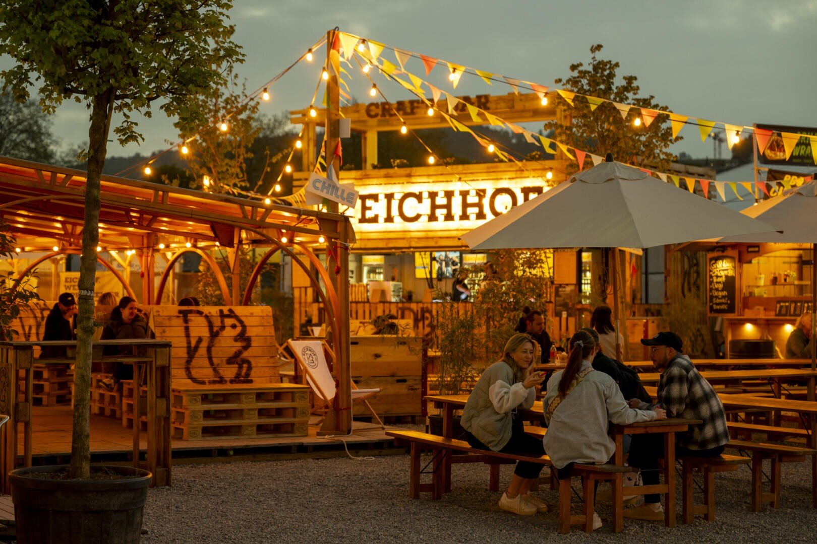 Abends sitzen die Menschen an Picknicktischen unter Lichterketten und Sonnenschirmen in einem Biergarten im Freien. Der warme Schein der Lichter und der Beschilderung schafft eine gemütliche Atmosphäre, und Bäume und Holzkonstruktionen umgeben den Sitzbereich.