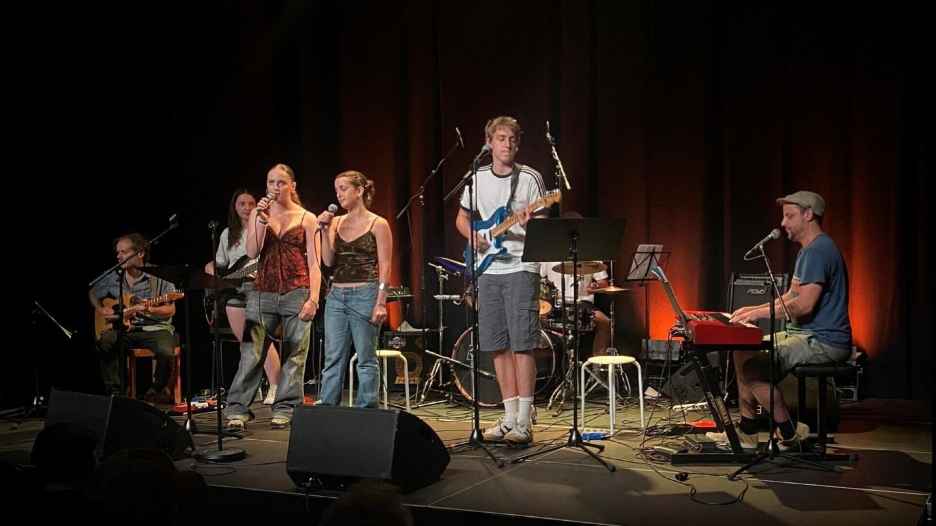 Auf der Bühne tritt eine Band mit sieben Mitgliedern auf, die Gitarre, Keyboard und Schlagzeug spielen und in Mikrofone singen, bei warmer Bühnenbeleuchtung und dunklem Hintergrund.