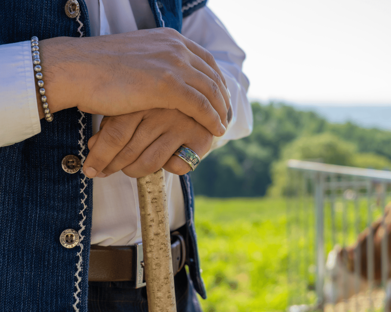 Eine Person, die eine Jeansweste, ein weißes Hemd, ein Armband und einen türkisfarbenen Ring trägt, stützt sich mit den Händen auf einen Holzstab, während im unscharfen Hintergrund Grünpflanzen und ein Metallgeländer zu sehen sind.
