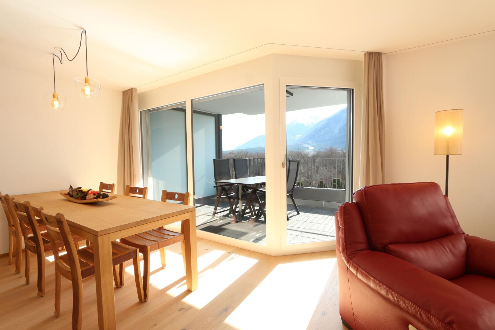 Ein heller Ess- und Wohnbereich mit einem Holztisch und sechs Stühlen, einem roten Ledersessel, einer hohen Stehlampe und großen Glasschiebetüren, die auf einen Balkon mit Bergblick führen.