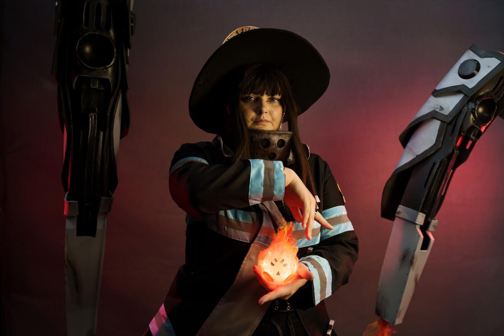 Eine Person in einem dunklen Kostüm mit blauen Streifen und einem großen Hut posiert mit den Händen um einen glühenden, feurigen Gegenstand. Zwei große mechanische Requisiten stehen auf beiden Seiten vor einem dunklen Hintergrund.