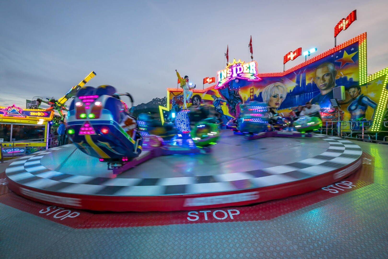 Ein buntes Fahrgeschäft im Vergnügungspark dreht sich mit unscharfen Bewegungen und blinkenden Lichtern vor einem düsteren Himmel. Lebhafte Kunstwerke und Leuchtreklamen schmücken das Fahrgeschäft, während STOP mehrfach auf die rote Plattformkante gedruckt ist.