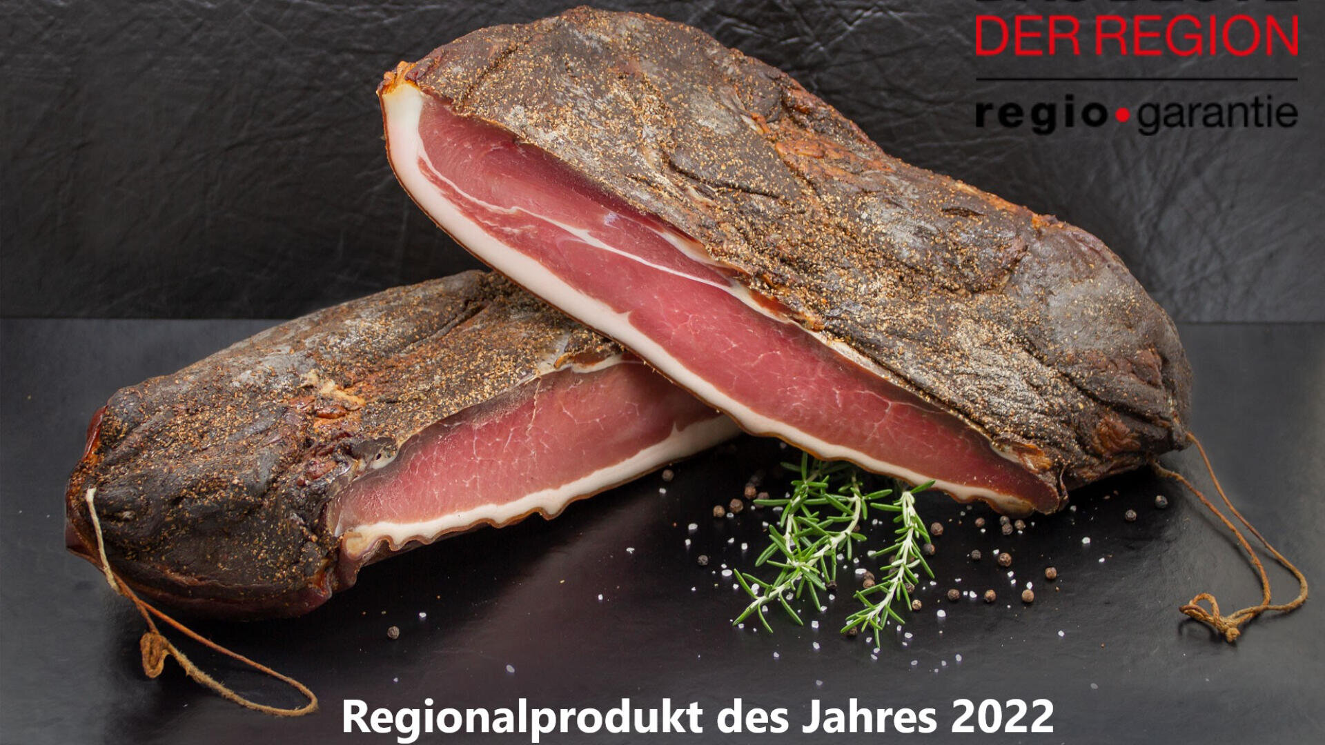 Zwei Stücke gepökeltes Fleisch auf einem schwarzen Untergrund mit Rosmarin und Pfefferkörnern. Der Text lautet auf Deutsch: „DER REGION regio.garantie“ und „Regionalprodukt des Jahres 2022“.