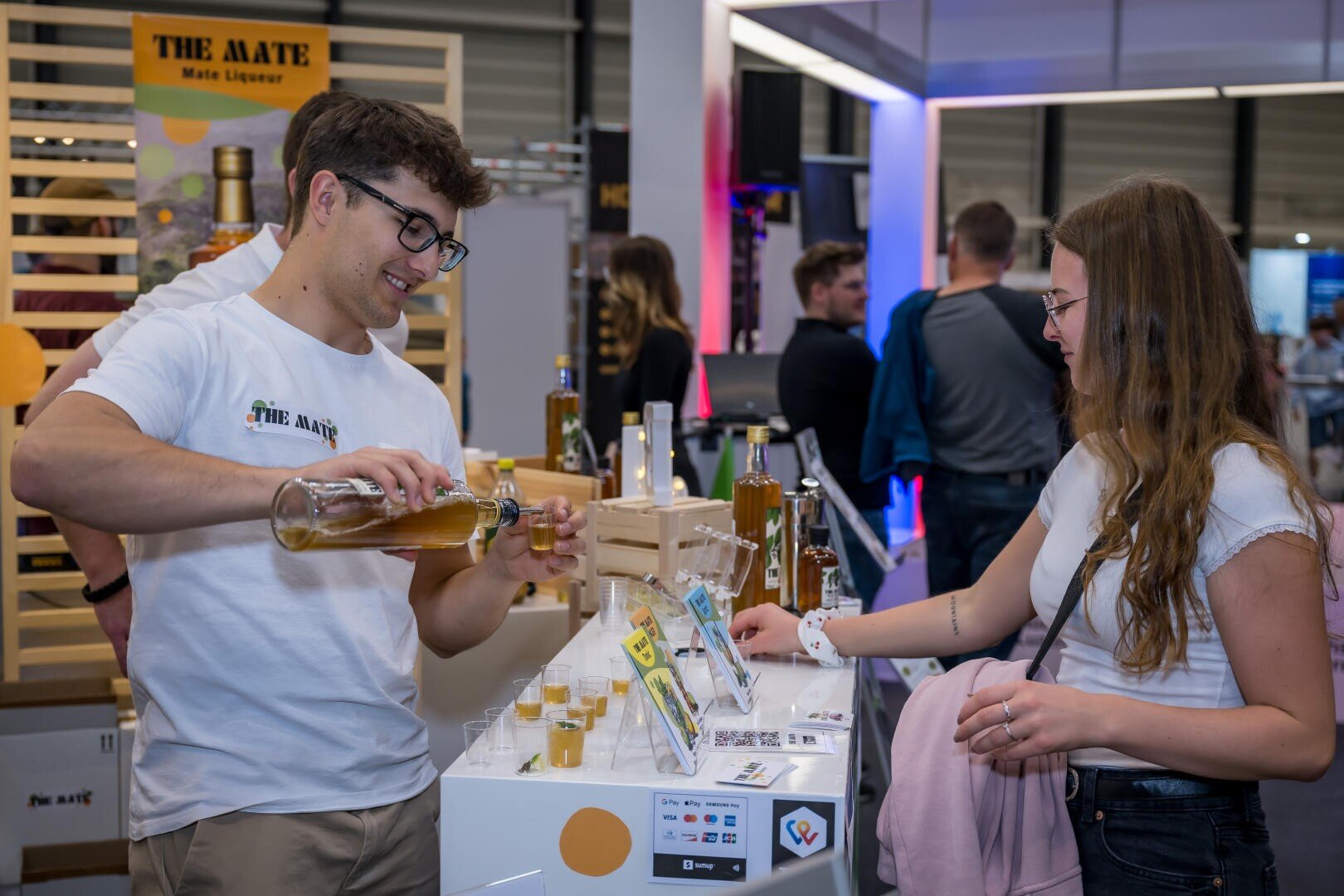 Ein Mann in einem weißen T-Shirt schenkt an einem Stand ein gelbes Getränk in kleine Becher ein, während eine Frau mit langen Haaren und Brille lächelnd zusieht. Auf dem Tisch stehen Flaschen und Display-Karten, im Hintergrund sind weitere Personen zu sehen.