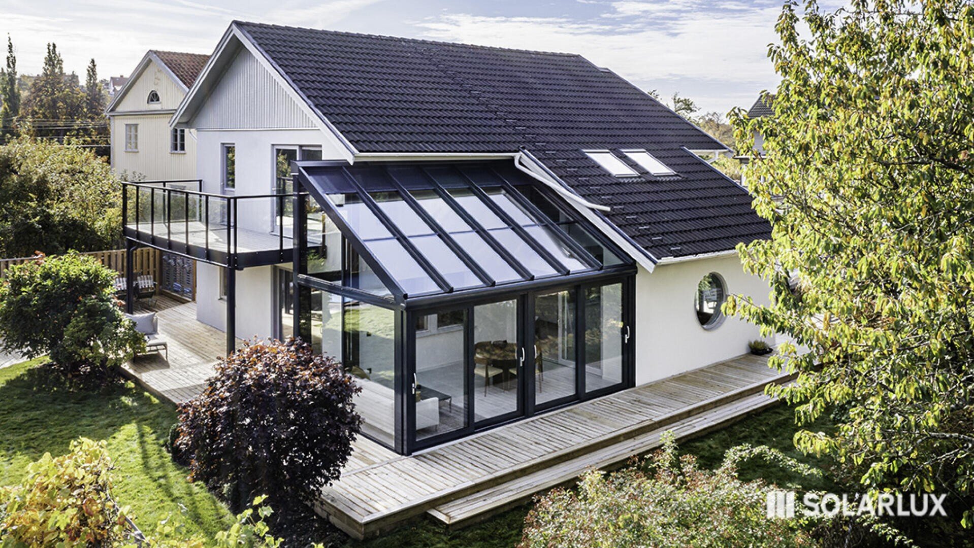 Ein modernes zweistöckiges Haus mit einem dunklen Dach und einem großen Glasanbau steht auf einer Holzterrasse, umgeben von üppigem Grün. Das Logo Solarlux erscheint in der unteren rechten Ecke.