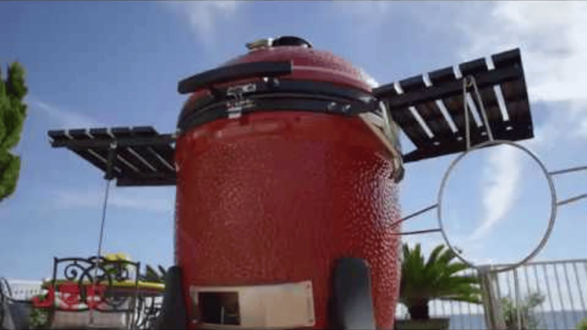 Ein großer roter Keramik-Außengrill mit schwarzen Seitenablagen steht auf einer Terrasse mit Metallmöbeln und grünen Pflanzen, die im Hintergrund unter einem blauen Himmel zu sehen sind.