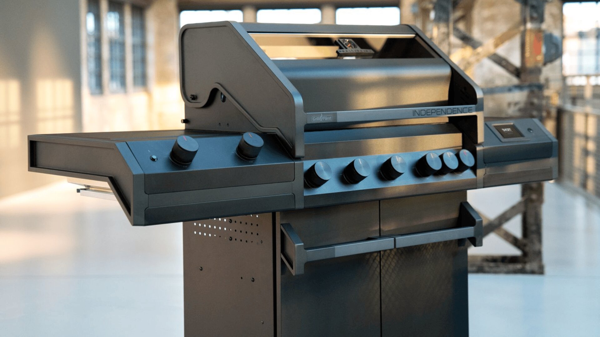 Ein moderner schwarzer Außengrill mit mehreren Drehreglern und einer Stahloberfläche, der im Innenbereich in der Nähe großer Fenster und freiliegender Metallträger aufgestellt ist.