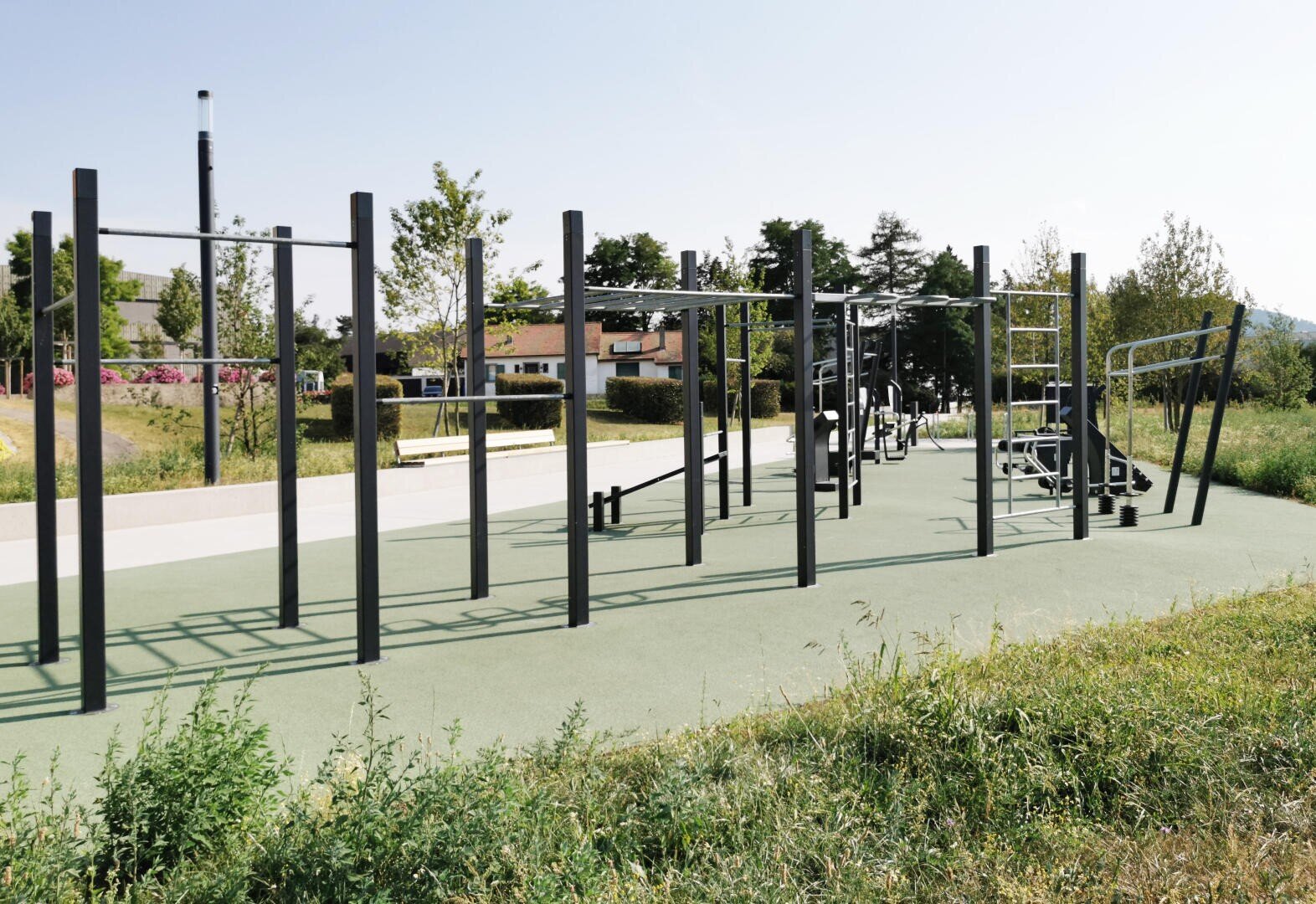 Ein Outdoor-Fitnesspark mit verschiedenen Trainingsgeräten, darunter Klimmzugstangen, Parallelbarren und Klettergerüste, umgeben von Gras, Bäumen und nahe gelegenen Gebäuden an einem sonnigen Tag.