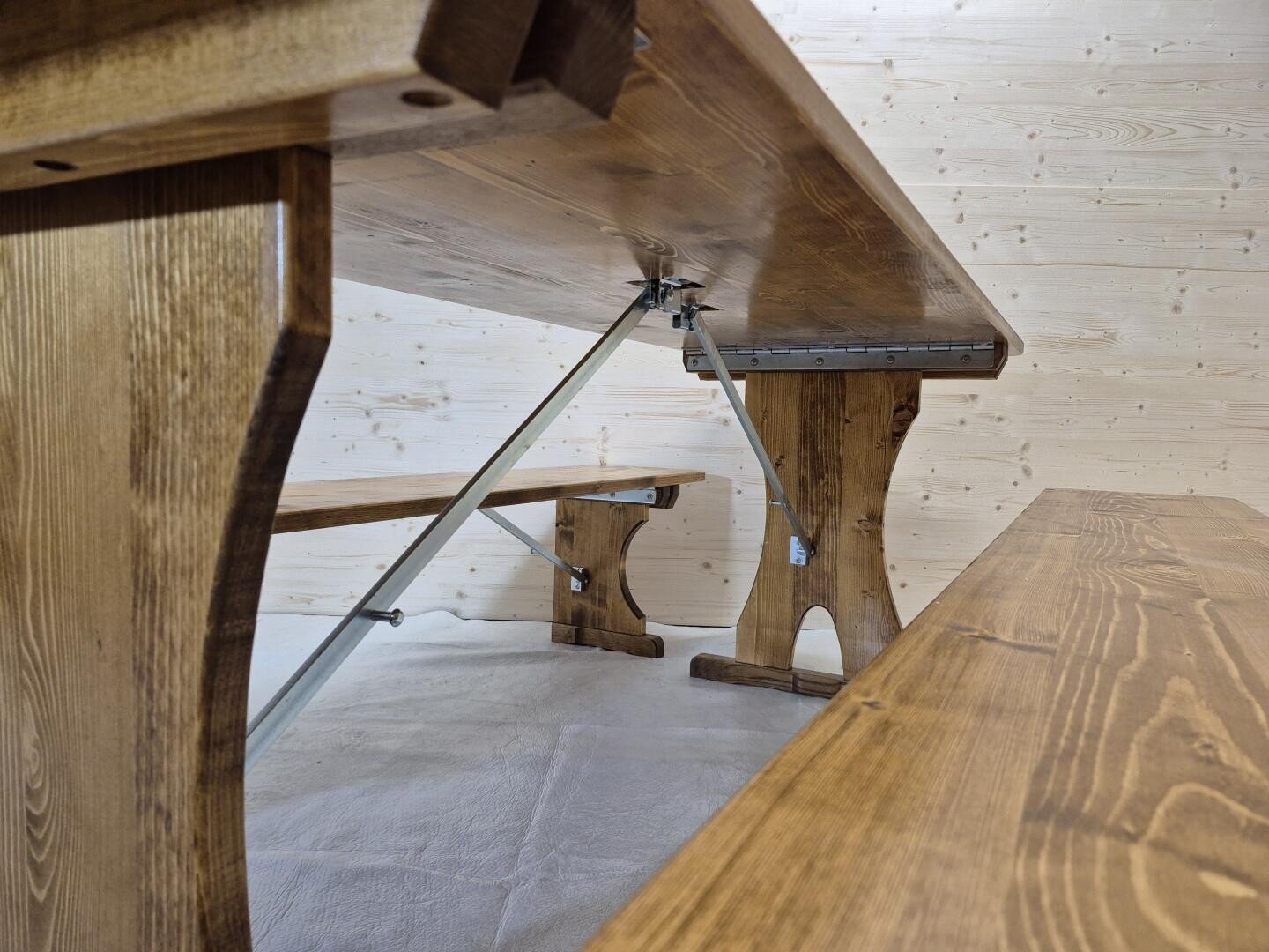 Blick aus geringer Höhe auf einen Holztisch und Bänke mit Metallstangen, die auf einem hellen Boden vor einer hellen holzgetäfelten Wand stehen. Die Möbel haben ein rustikales, robustes Aussehen.