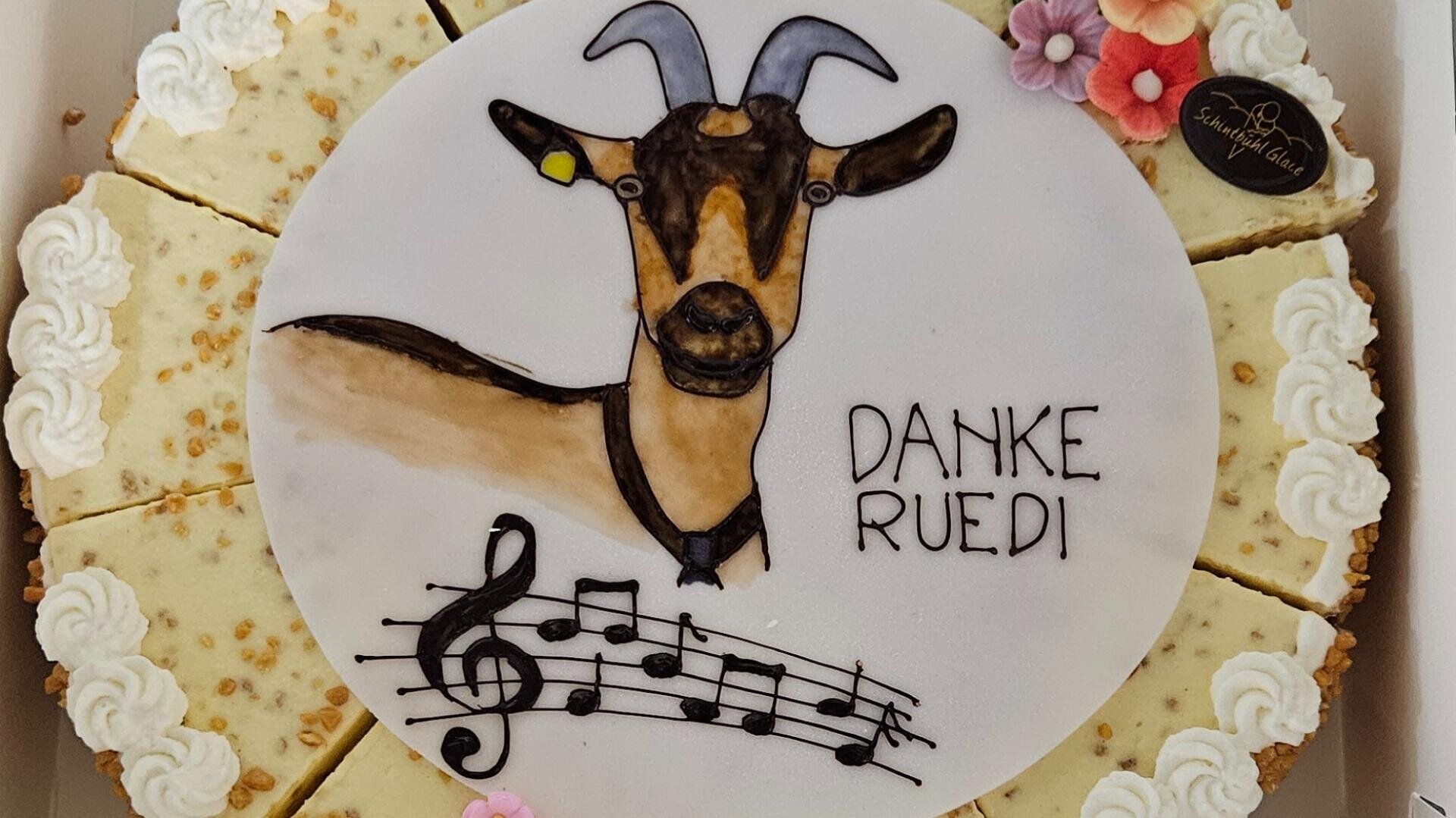 Eine runde Torte, verziert mit der Zeichnung einer Ziege, Musiknoten und der Aufschrift DANKE RUEDI. Die Torte wird in Scheiben geschnitten und mit Sahne und kleinen rosa und weißen Fondantblumen gekrönt.