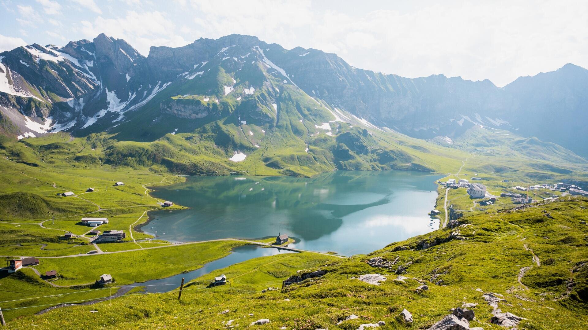 Ein klarer Bergsee spiegelt die umliegenden grünen Hügel und schroffen, schneebedeckten Gipfel wider; verstreute kleine Gebäude und verschlungene Wege durchziehen die lebendige, sonnenbeschienene Landschaft unter einem teilweise bewölkten Himmel.