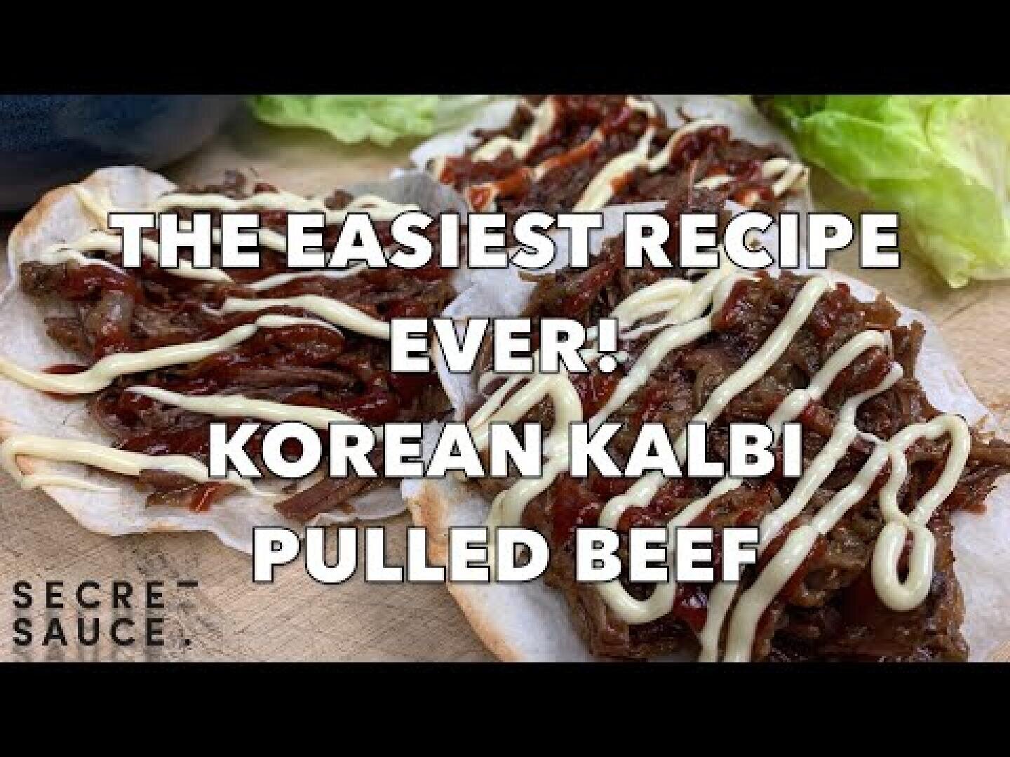 Zwei Stücke Brot, belegt mit zerkleinertem koreanischem Kalbi Pulled Beef und einem Spritzer Soße, mit Salat im Hintergrund. Der überlagerte Text lautet: Das einfachste Rezept aller Zeiten! Koreanisches Kalbi Pulled Beef.