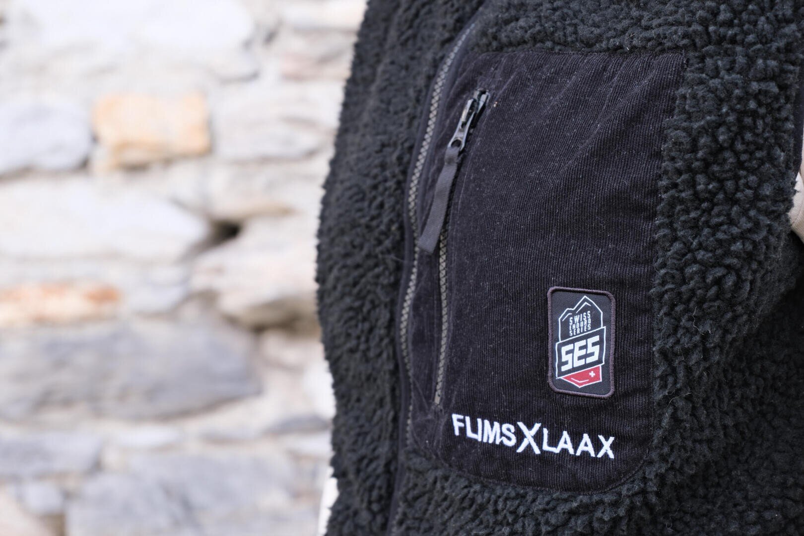 Nahaufnahme einer schwarzen Fleece-Weste mit einer Brusttasche mit Reißverschluss, auf der die Logos SES und FLIMS X LAAX vor einem unscharfen Hintergrund aus Steinmauern aufgestickt sind.