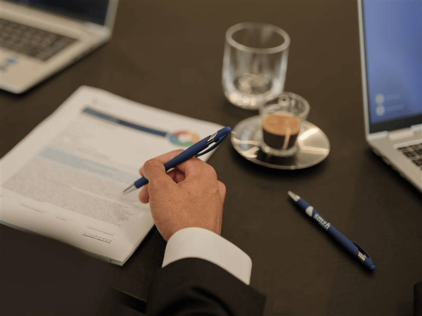 Eine Person im Anzug hält einen Stift über ein Dokument an einem Schreibtisch, in dessen Nähe ein Laptop, ein Glas Wasser, eine Tasse Espresso auf einer Untertasse und ein weiterer Stift zu sehen sind.