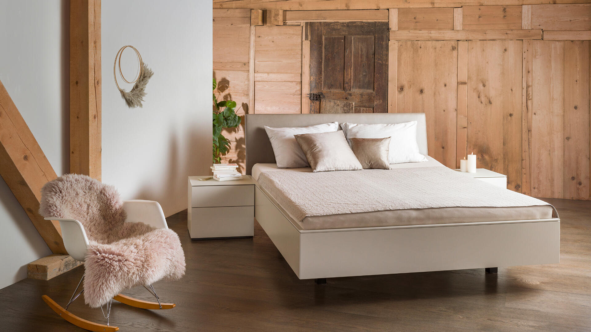 Ein minimalistisches Schlafzimmer mit einem weißen Bett, beiger Bettwäsche und zwei Nachttischen. Im Hintergrund sind eine Holzwand und eine Holztür zu sehen. Im Vordergrund steht ein weißer Schaukelstuhl mit einem rosa, flauschigen Überzug. Kerzen und Deko-Elemente sorgen für eine gemütliche Atmosphäre.