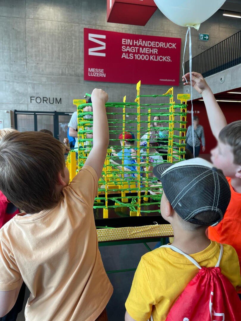 Drei Kinder interagieren mit einer großen, farbenfrohen Murmelbahn in einem Innenraum. Im Hintergrund ist eine Betonwand mit roter Beschriftung in deutscher Sprache und den Worten MESSE LUZERN und FORUM zu sehen.