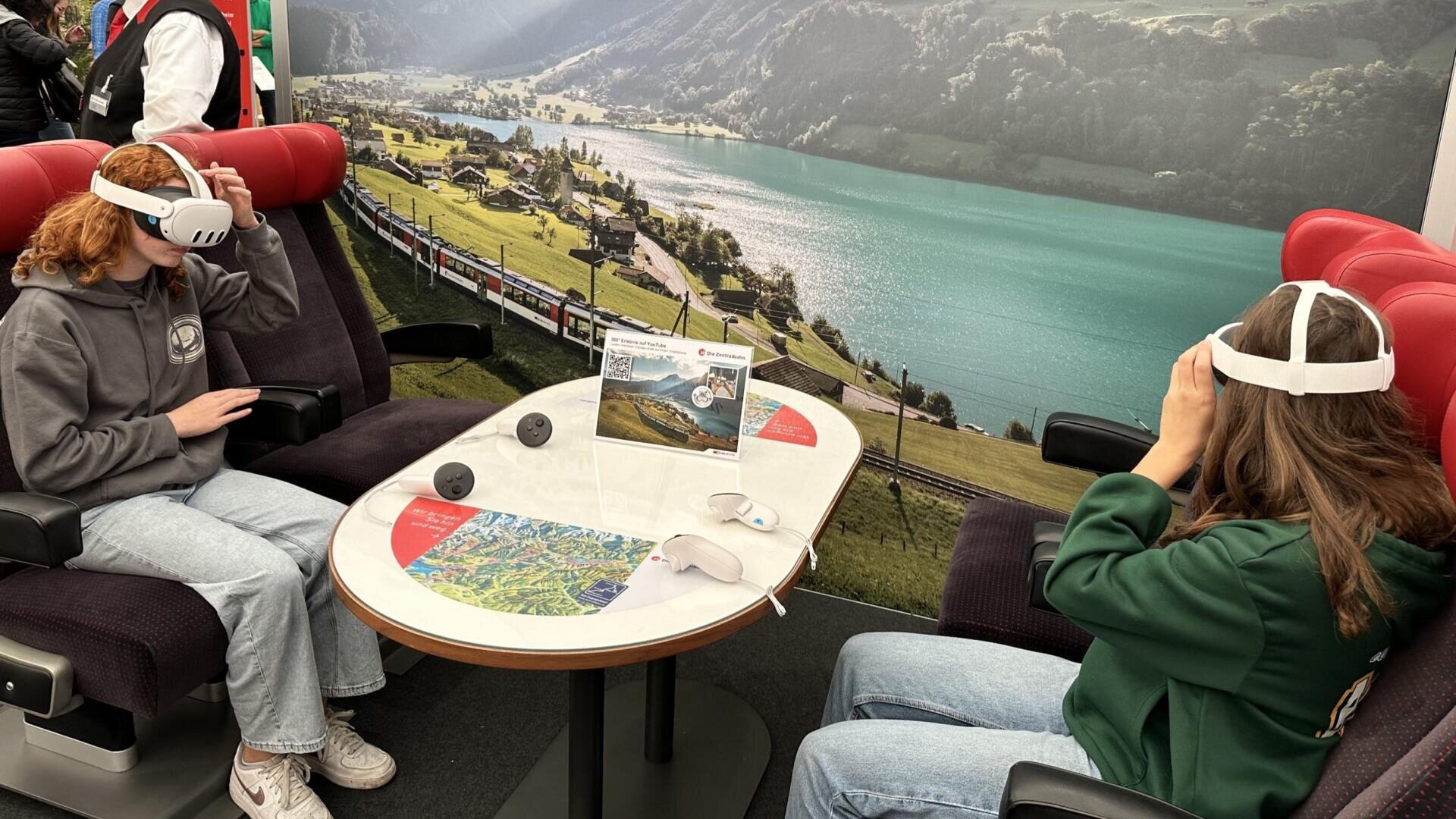 Zwei Personen, die VR-Headsets tragen, sitzen sich in roten Zugsitzen an einem kleinen Tisch gegenüber. Hinter ihnen ist ein malerischer Schweizer Alpensee und ein Eisenbahn-Wandbild zu sehen.