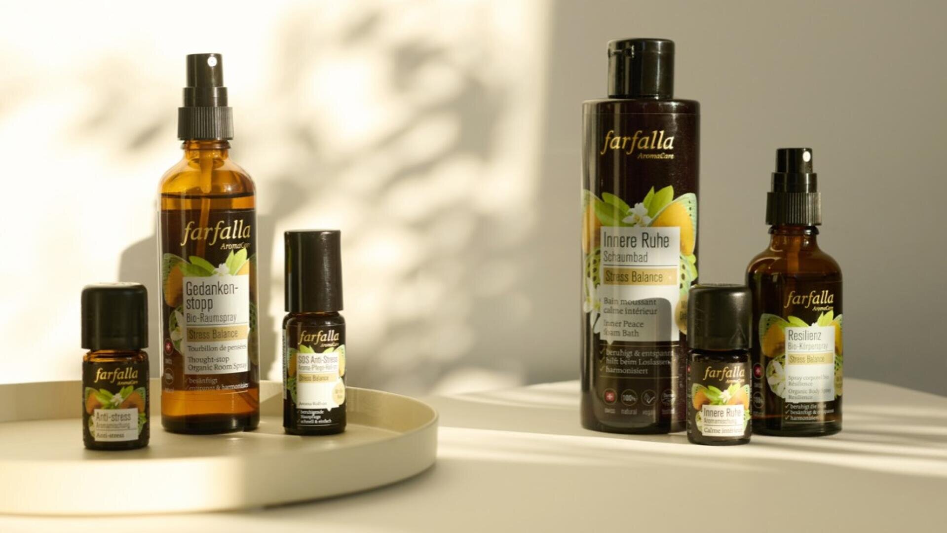Sechs ätherische Öl- und Wellnessprodukte der Marke Farfalla sind auf einer hellen Fläche angeordnet, einige davon auf einem runden Tablett, das von sanftem Sonnenlicht beleuchtet wird. Der Hintergrund weist Licht- und Schattenmuster auf.