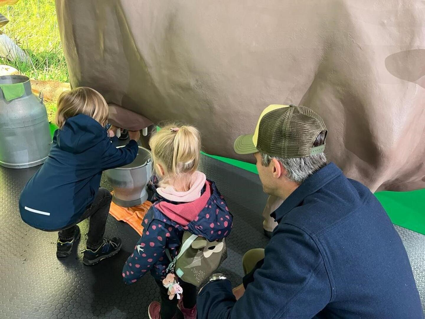 Zwei Kinder und ein Erwachsener knien neben einer Modellkuh, von der ein Kind so tut, als würde es sie in einen Metalleimer melken. Der Erwachsene schaut zu, während die Kinder Jacken tragen und die Szene lehrreich wirkt.