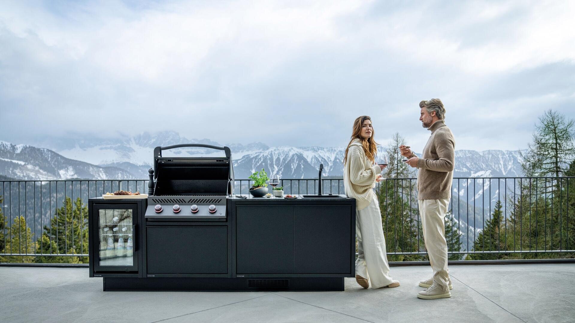 Ein Mann und eine Frau stehen auf einer Terrasse neben einer modernen Grillküche im Freien, unterhalten sich und halten Getränke in der Hand, während im Hintergrund schneebedeckte Berge und Bäume zu sehen sind.
