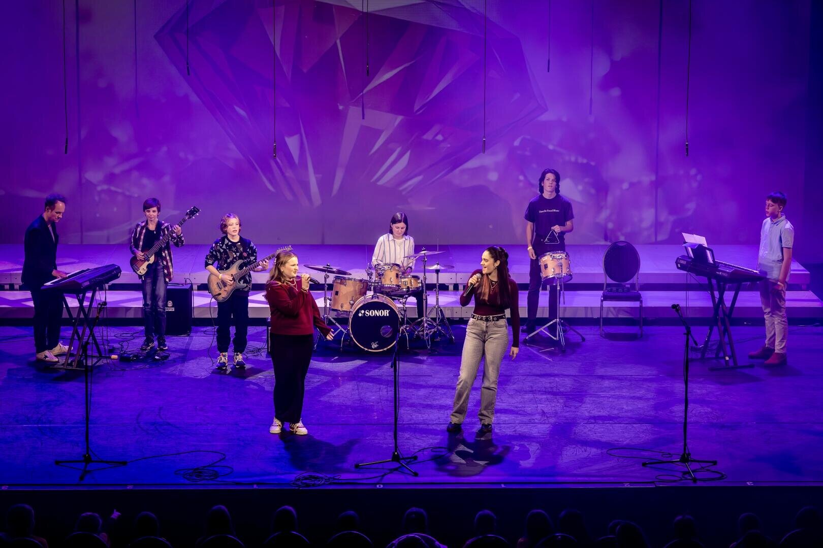 Eine Band mit zwei Sängern, einem Schlagzeuger, zwei Keyboardern, einem Gitarristen, einem Bassisten und einem Schlagzeuger, mit Mikrofonen und einem geometrischen Hintergrund hinter ihnen, tritt auf der Bühne unter lila Beleuchtung auf.