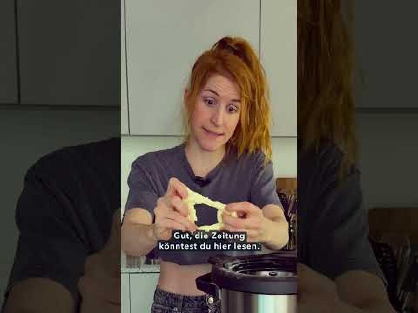 Eine rothaarige Frau steht in einer Küche und hält mit überraschtem Blick ein Stück Käse über einen Elektroherd. Die deutschen Untertitel am unteren Rand lauten: "Gut, die Zeitung könntest du hier lesen.