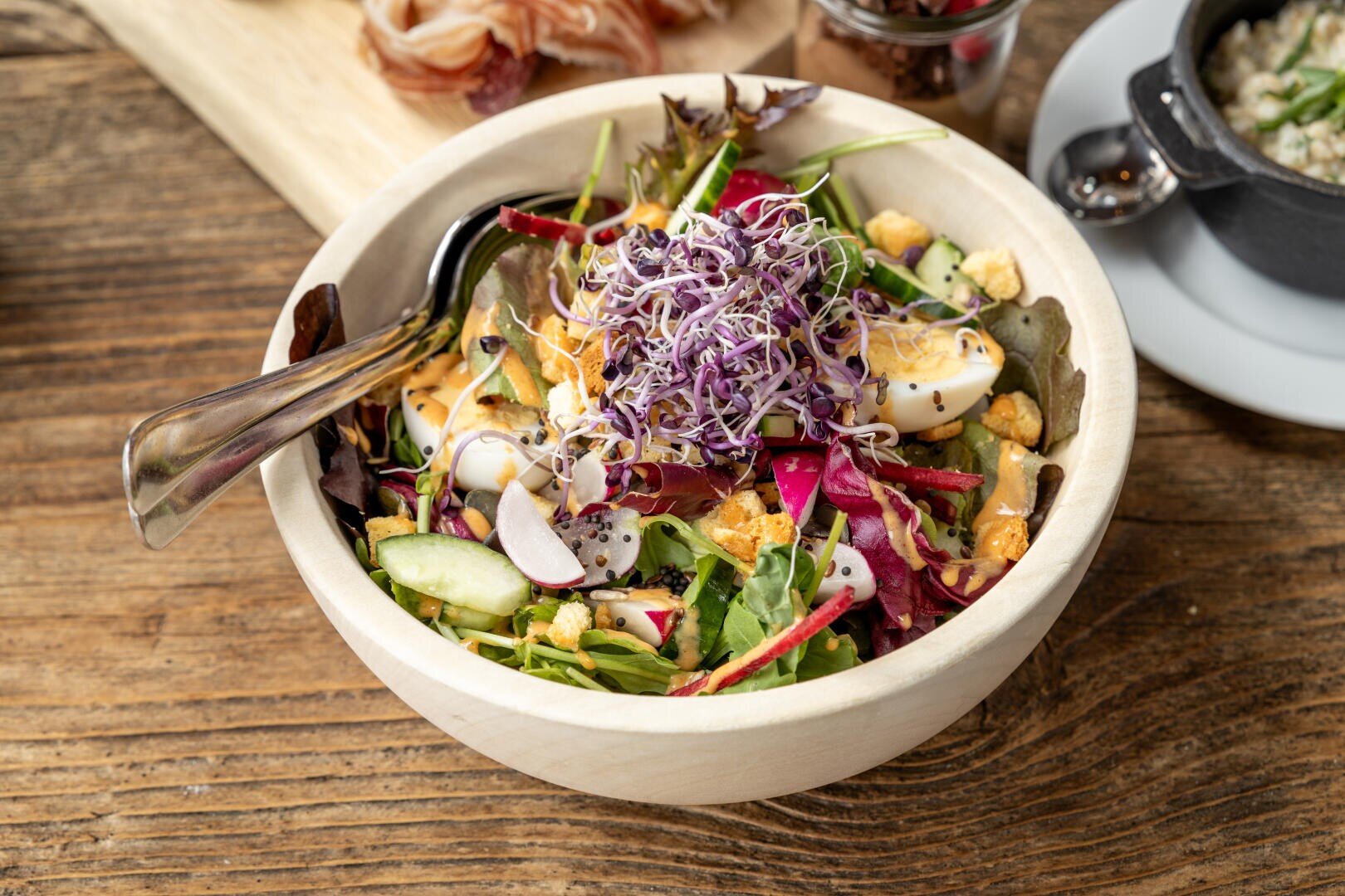 Eine Holzschüssel, gefüllt mit gemischtem Salat, Radieschenscheiben, Gurken, hart gekochten Eierscheiben, Croutons und einem Haufen Alfalfasprossen, mit einer Gabel darin, auf einem rustikalen Holztisch stehend.