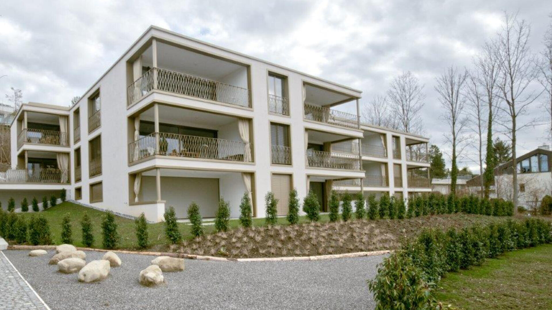 Ein modernes, dreistöckiges Wohnhaus mit großen Balkonen, umgeben von Grünanlagen, Sträuchern und einem Kiesweg unter einem bewölkten Himmel.