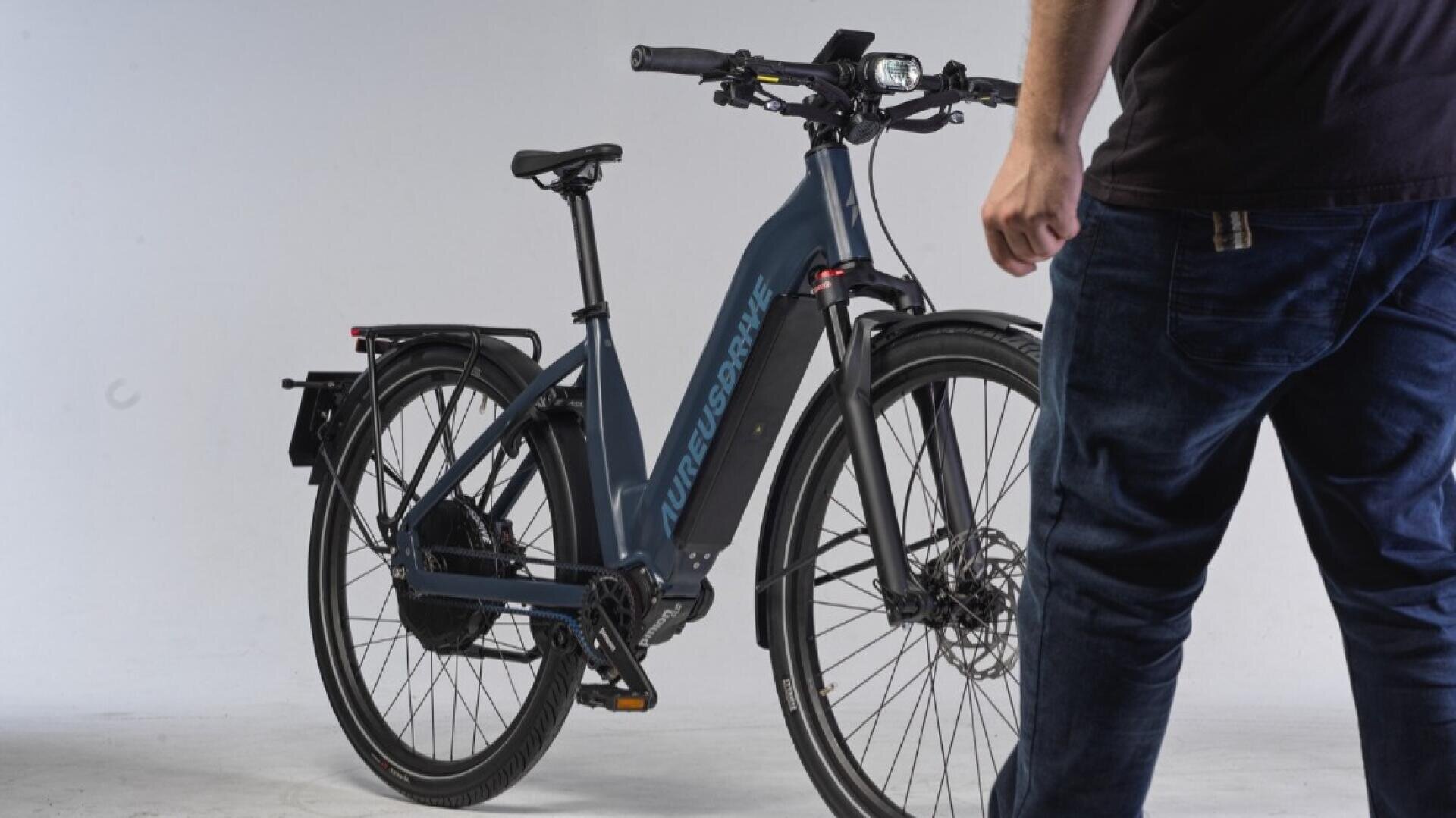 Eine dunkel gekleidete Person steht neben einem dunkelblauen E-Bike mit der Aufschrift PURE URBAN auf dem Rahmen in einem Studio mit weißem Hintergrund.