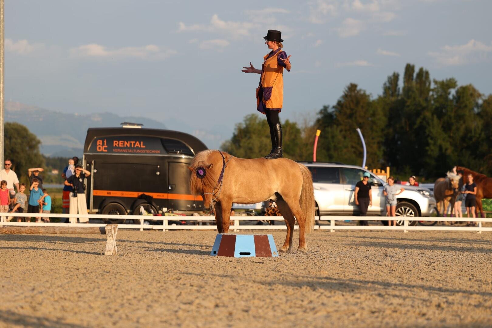 Ein Darsteller mit Zylinder und orangefarbenem Outfit steht auf dem Rücken eines kleinen, hellbraunen Pferdes mit einer Schleife, während einer Veranstaltung im Freien mit Zuschauern und Fahrzeugen im Hintergrund.