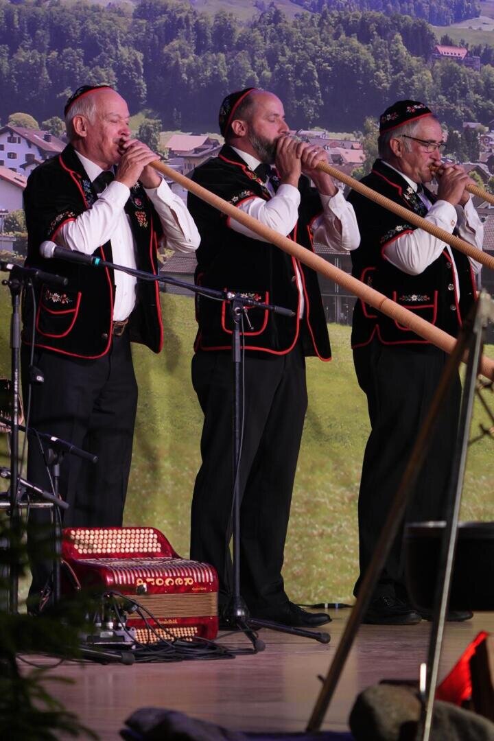 Drei Männer in traditioneller Schweizer Kleidung spielen auf der Bühne lange hölzerne Alphörner vor einer Kulisse aus grünen Hügeln und Häusern. Ein Akkordeon liegt in der Nähe auf dem Boden.