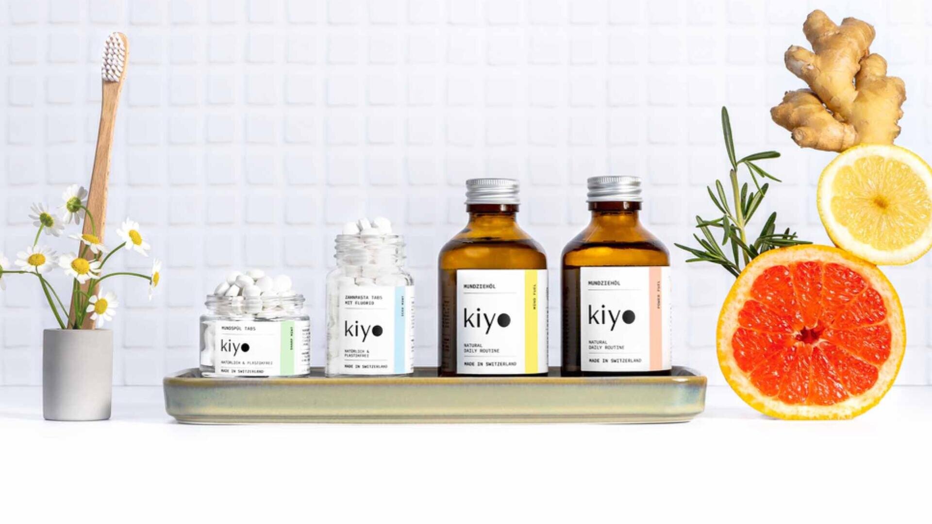 Auf einem Tablett stehen Flaschen und Gläser mit Kiyo-Nahrungsergänzungsmitteln, umgeben von einer Zahnbürste in einer Vase, Kamillenblüten, Ingwer, Zitrone, Grapefruithälften und Rosmarin, vor einem weiß gekachelten Hintergrund.