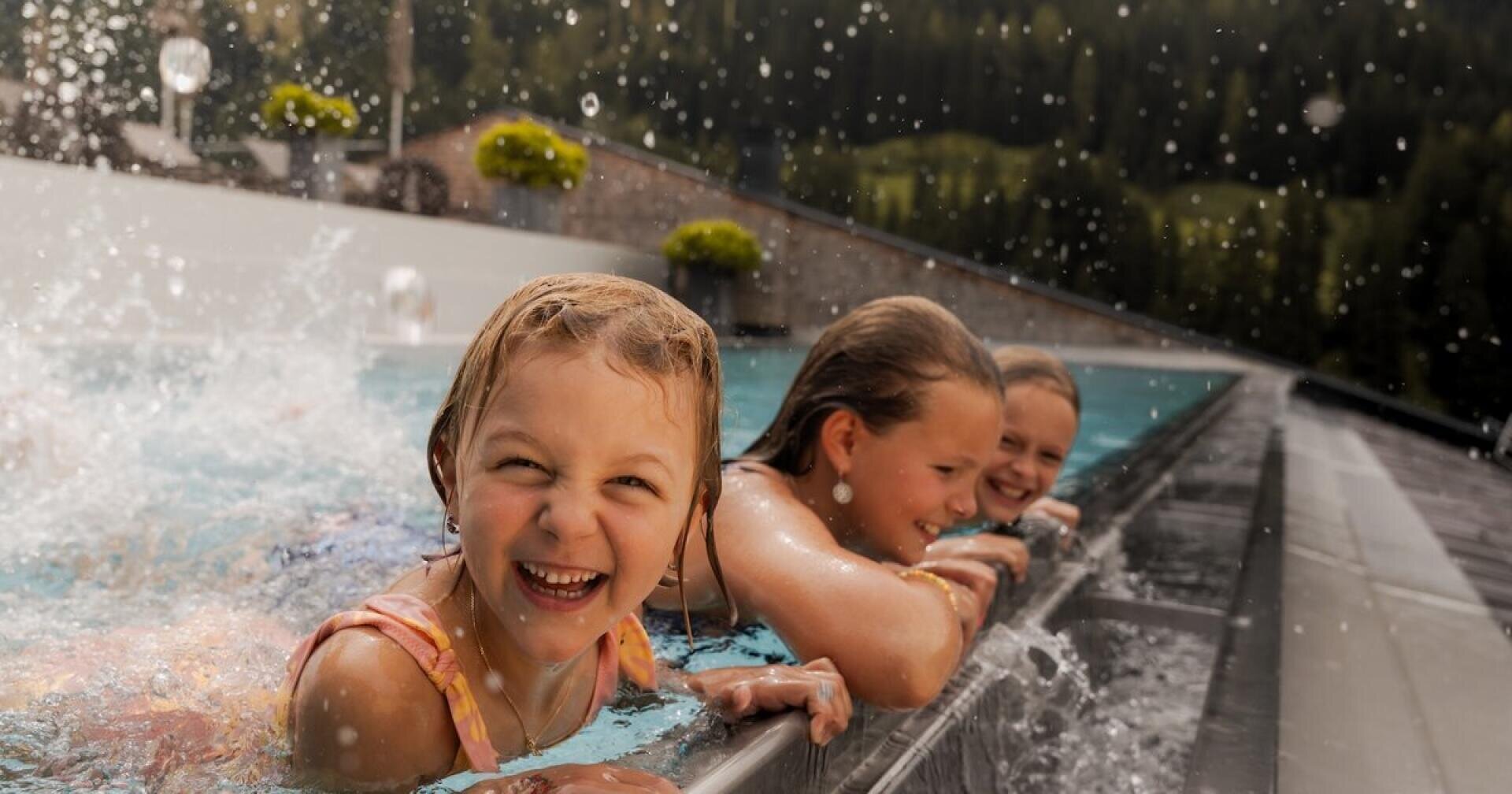 Drei lächelnde Kinder liegen am Rande eines Schwimmbeckens und planschen und lachen miteinander. Wassertropfen liegen in der Luft, und im Hintergrund sind Bäume und die Landschaft im Freien zu sehen.