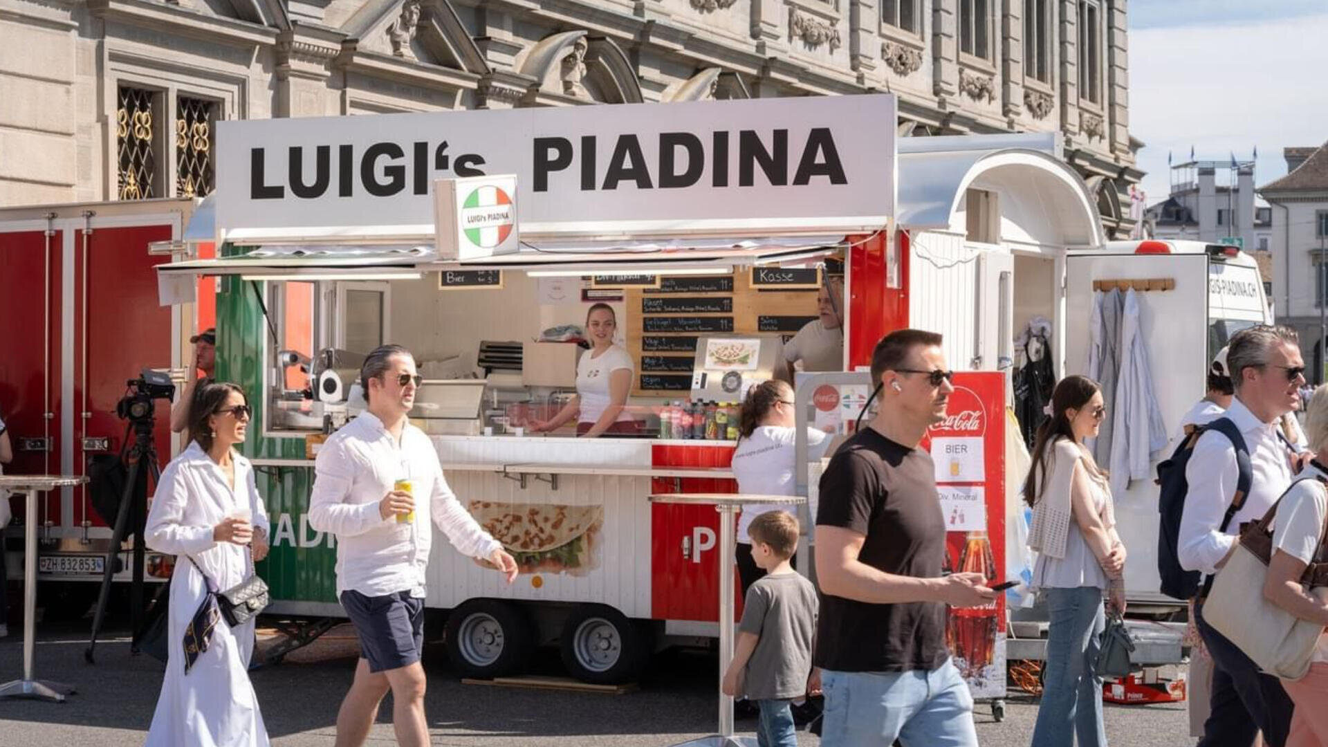 An einem sonnigen Tag kommen Passanten vorbei und bestellen bei „Luigi’s Piadina“, einem Foodtruck mit einem rot-weiß-grünen Schild, der auf einer Straße in der Stadt geparkt ist. Im Inneren des Trucks steht ein Kellner und lächelt.