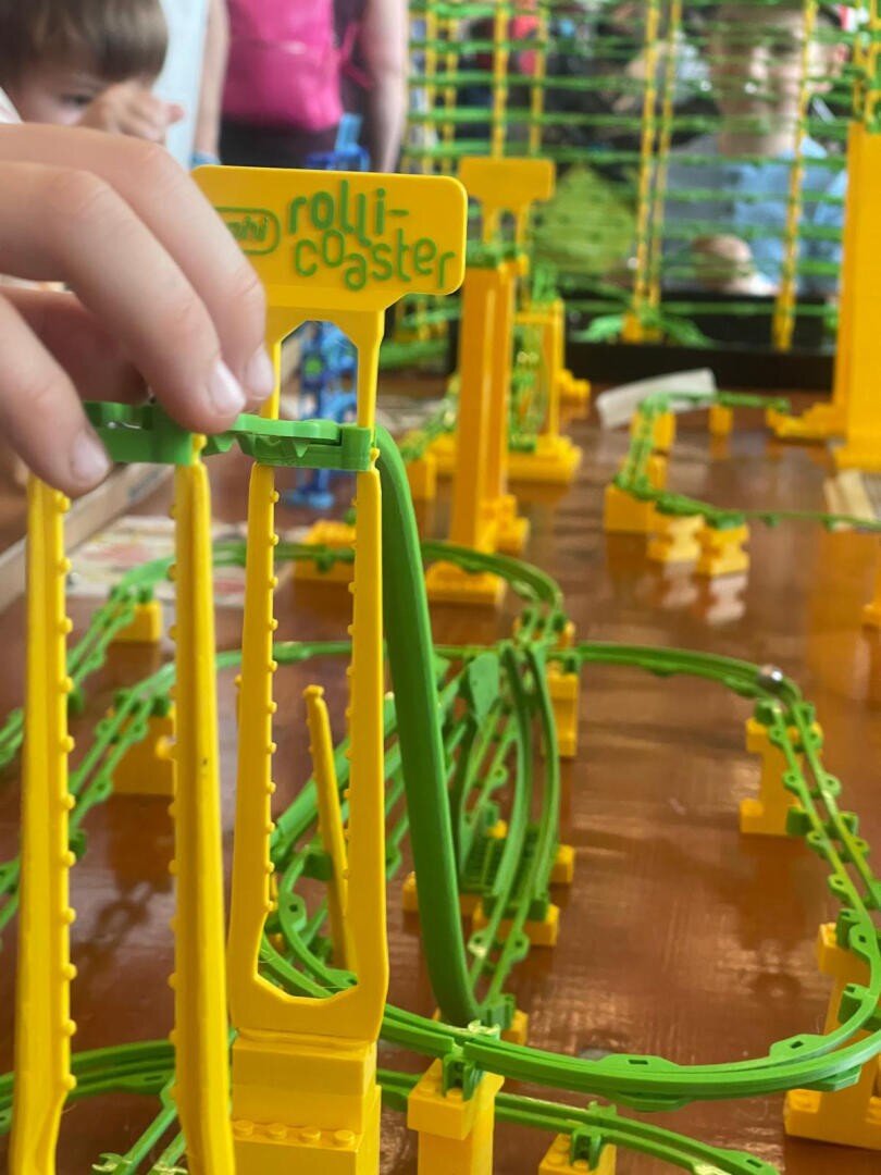 Eine Kinderhand interagiert mit einem gelben und grünen Spielzeug-Achterbahnset mit der Aufschrift rolli-coaster, wobei im Hintergrund Schienen und Strukturen zu sehen sind. Andere Kinder sind im Hintergrund verschwommen.