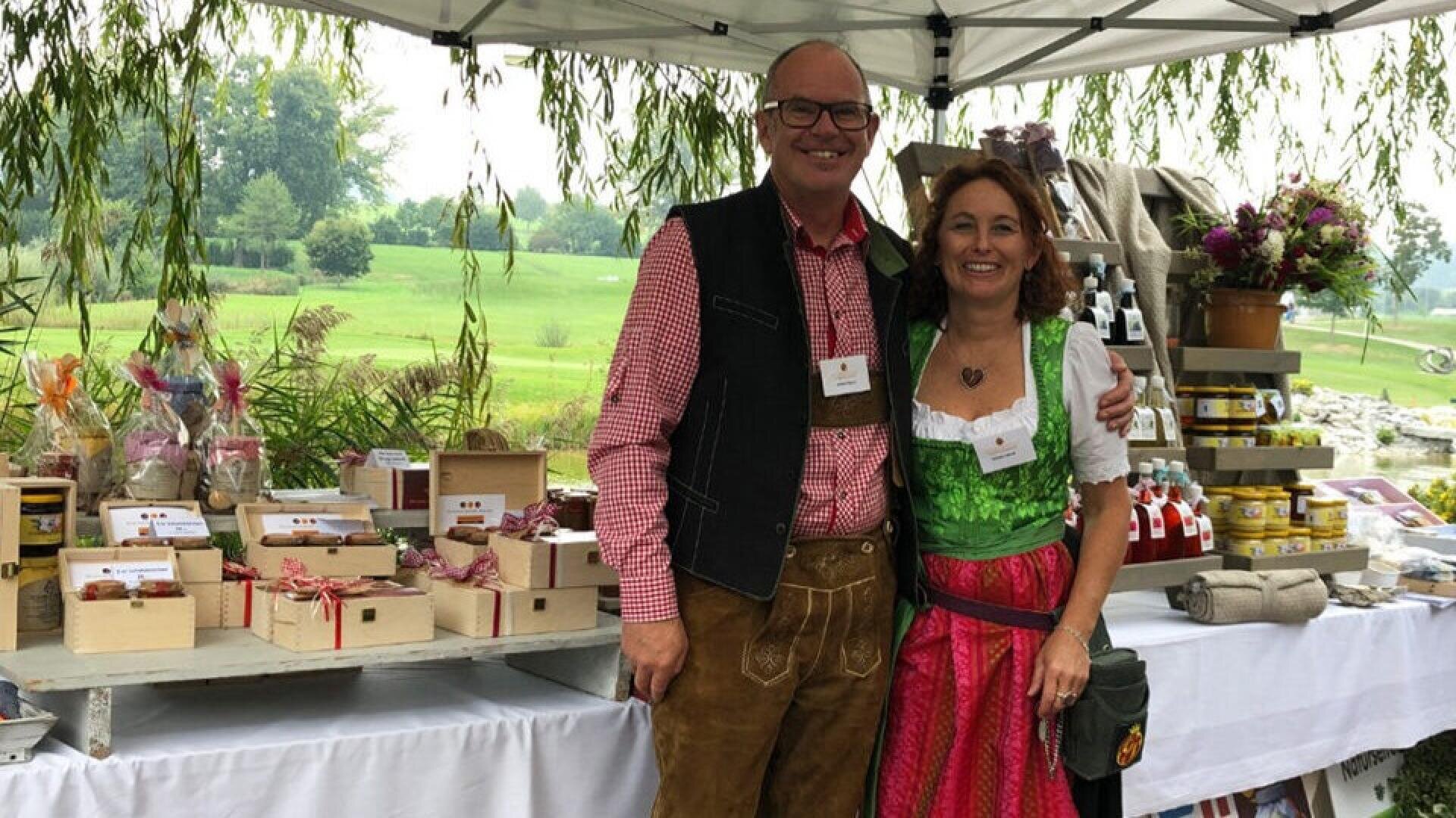 Ein lächelnder Mann und eine lächelnde Frau in traditioneller bayerischer Kleidung stehen unter einem Vordach an einem Marktstand im Freien mit Geschenkboxen, Gläsern und Flaschen auf Tischen. Im Hintergrund sind Bäume und Gras zu sehen.