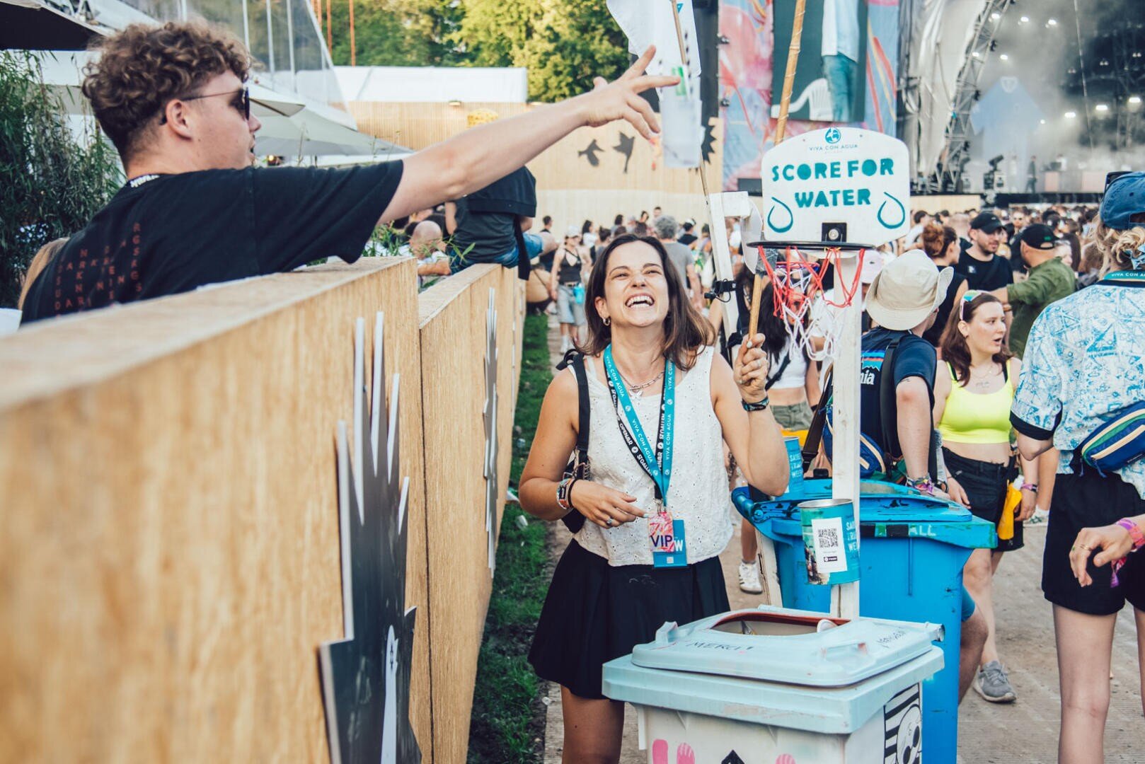 Eine Frau lächelt, während sie einen Basketballkorb mit der Aufschrift "Score for Water" hält, der an einem Mülleimer befestigt ist. Ein Mann wirft einen Ball in Richtung des Korbes auf einem Outdoor-Festival mit anderen Teilnehmern im Hintergrund.