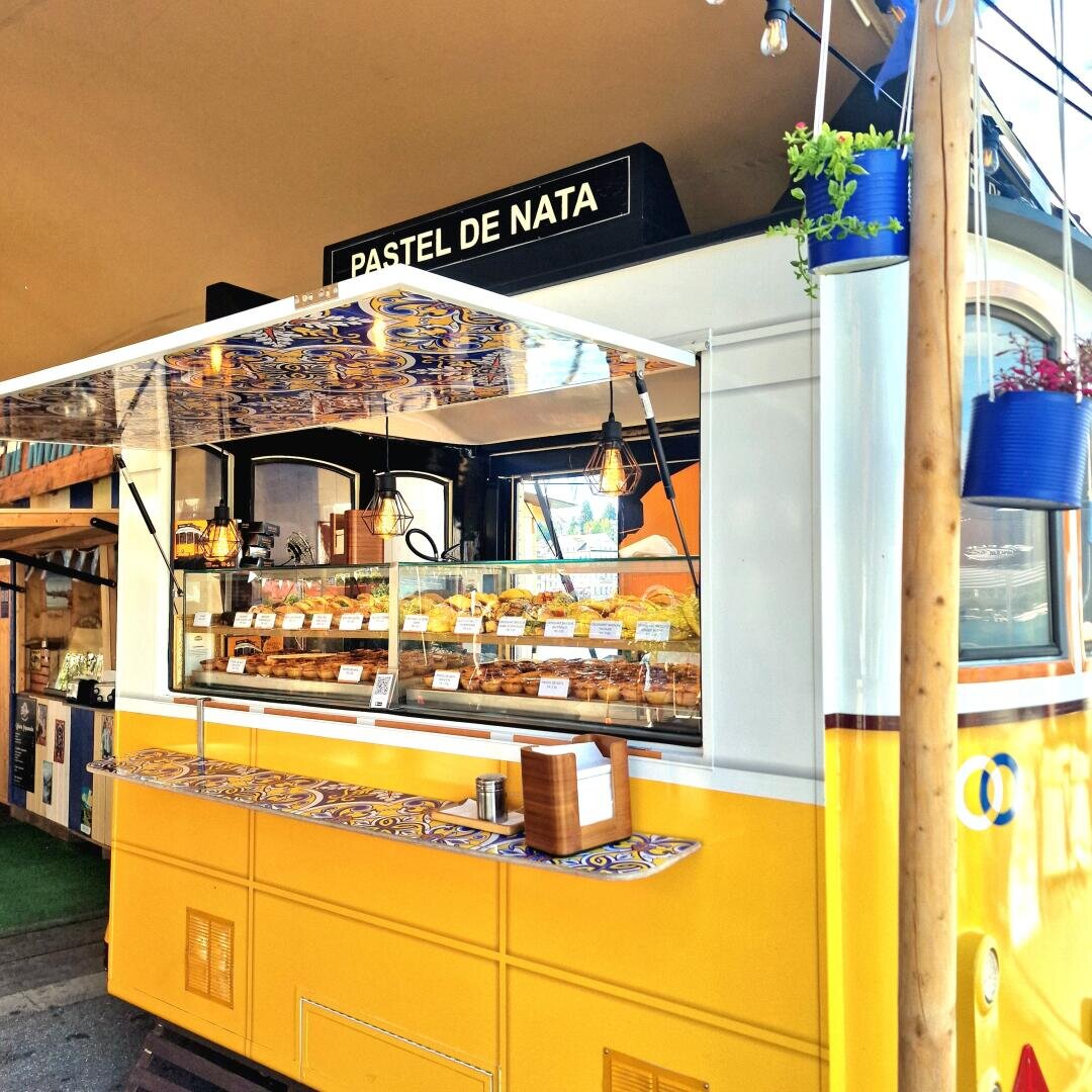 Ein gelb-weißer Imbisswagen mit der Aufschrift Pastel de Nata stellt Gebäck in einer Glasvitrine aus. Im Inneren hängen warme Lichter, und blaue Kacheln zieren die Theke. Blaue Blumentöpfe hängen an einem Holzpfosten in der Nähe.