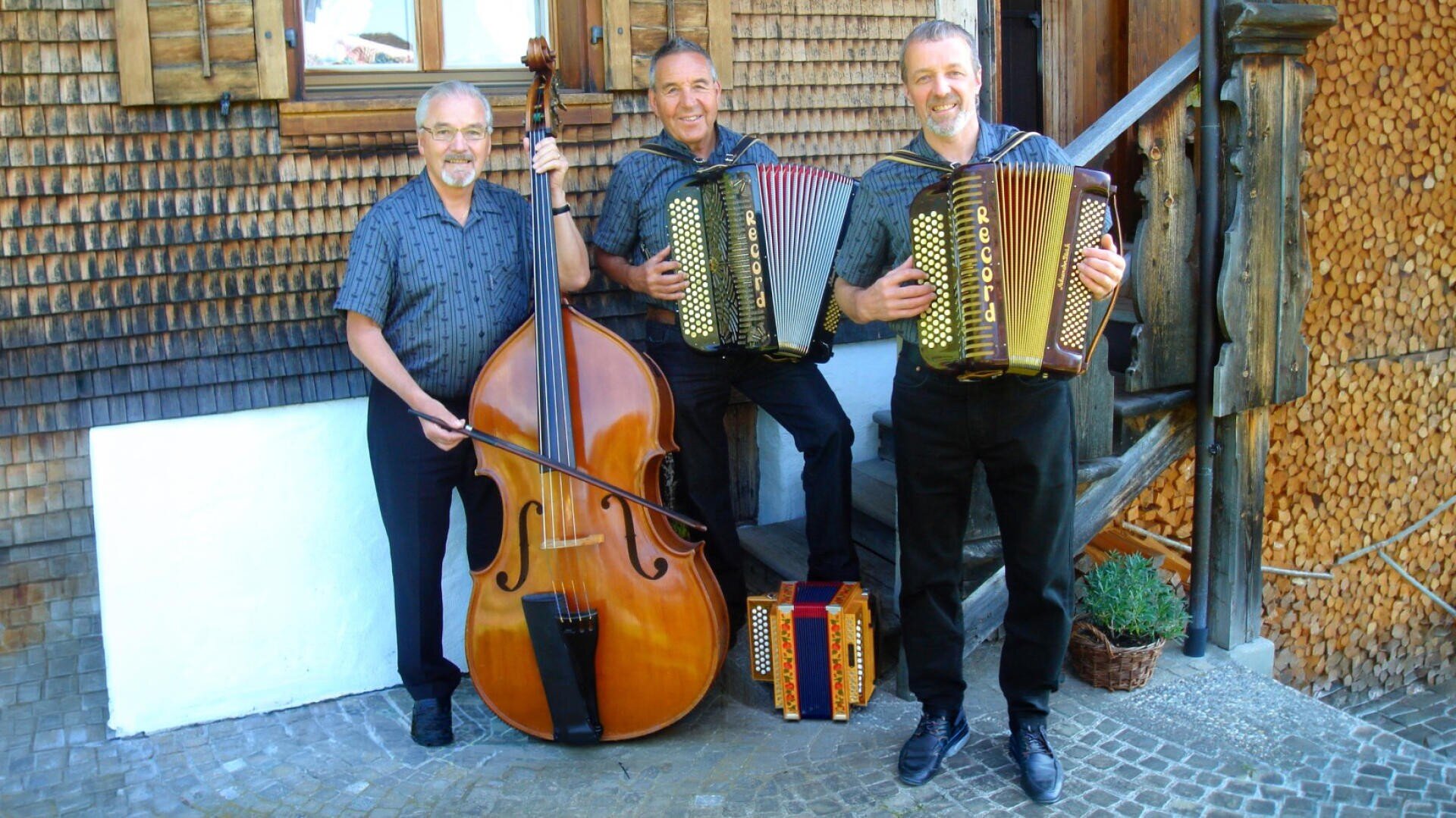 Drei lächelnde Männer, von denen zwei ein Akkordeon und einer einen Kontrabass in der Hand halten, posieren gemeinsam vor einem rustikalen Holzgebäude. Ein kleines Akkordeon liegt neben ihnen auf dem Boden.